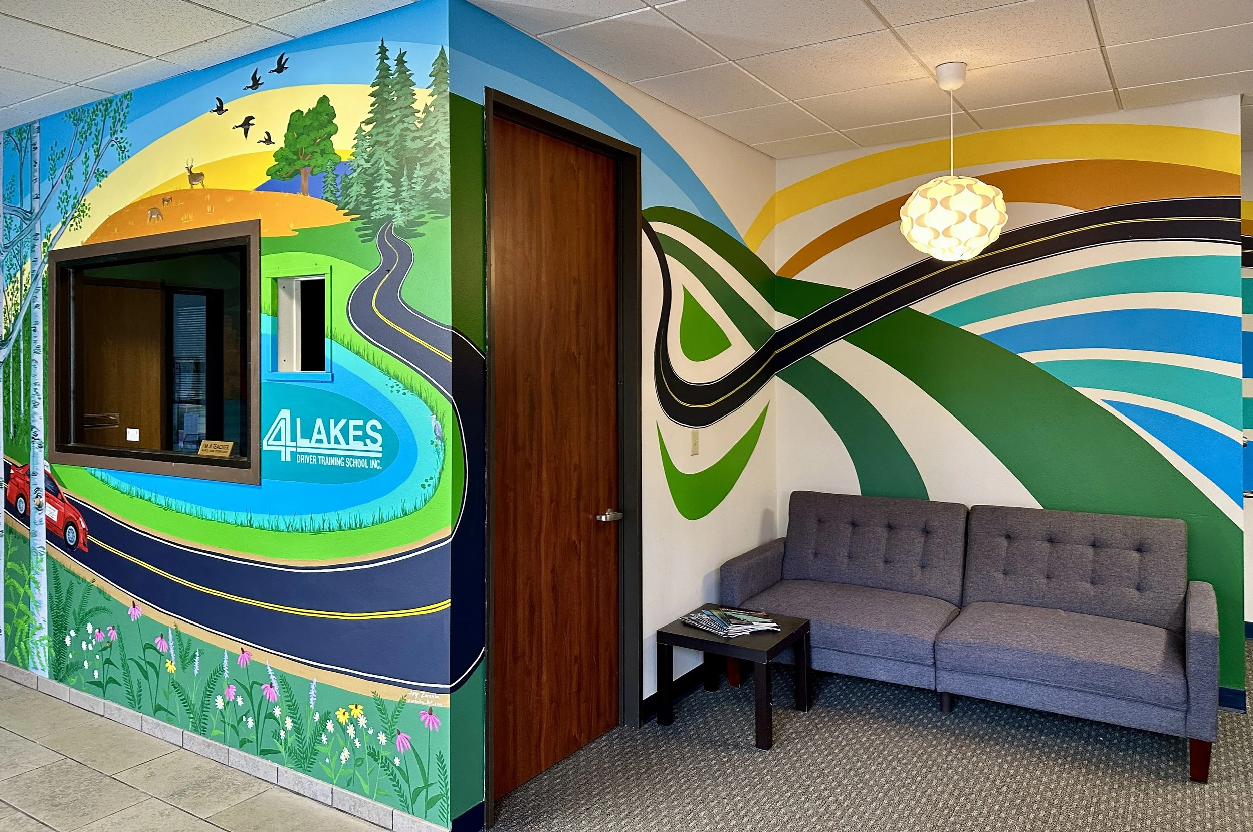 Colorful mural featuring a winding road, a lake with the text '4 Lakes Driver Training School Inc.', trees, and wildlife, with a gray couch, small table with magazines, dark wood door, window, and hanging light fixture in a waiting area.