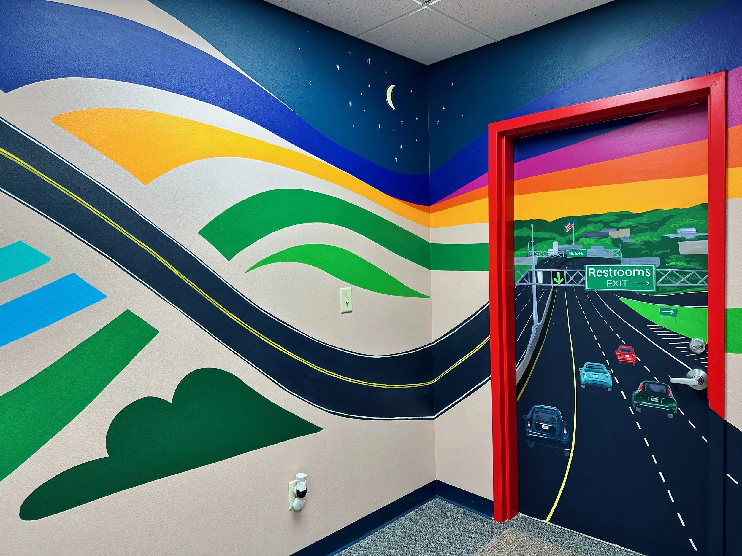 Colorful wall mural of a roadway with cars, green hills, trees, and a sunset sky, with a door painted as a highway exit sign indicating restrooms.