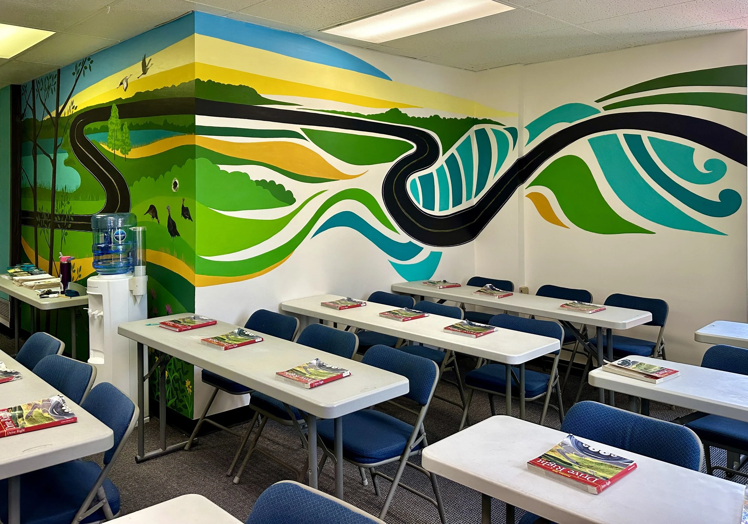 Classroom with painting of a winding road through green fields and hills on the wall, and desks with notebooks and chairs.