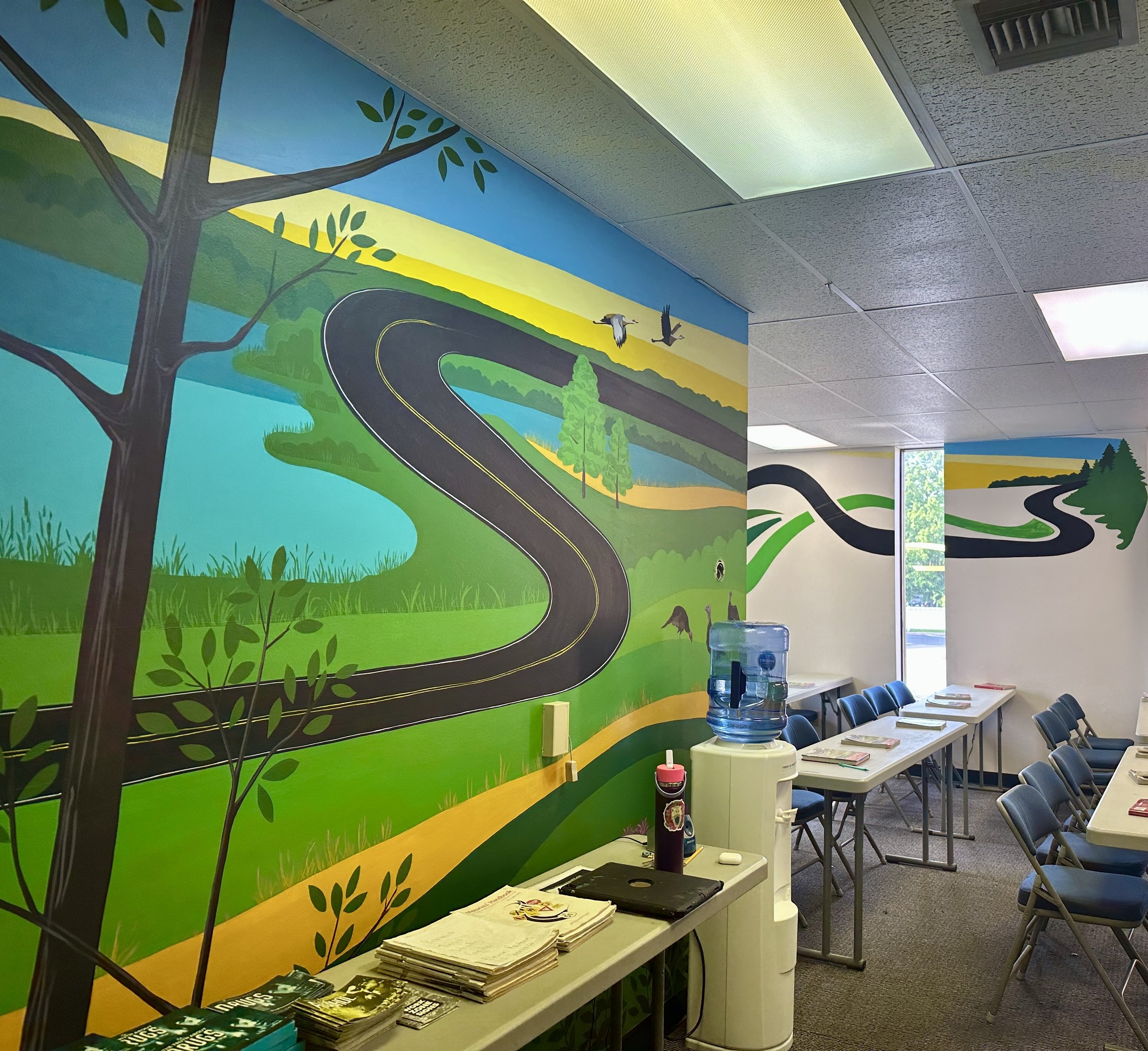 Interior of a room with a colorful mural of a winding road, trees, birds, and a lake painted on the wall, with tables, chairs, a water dispenser, and reading materials.