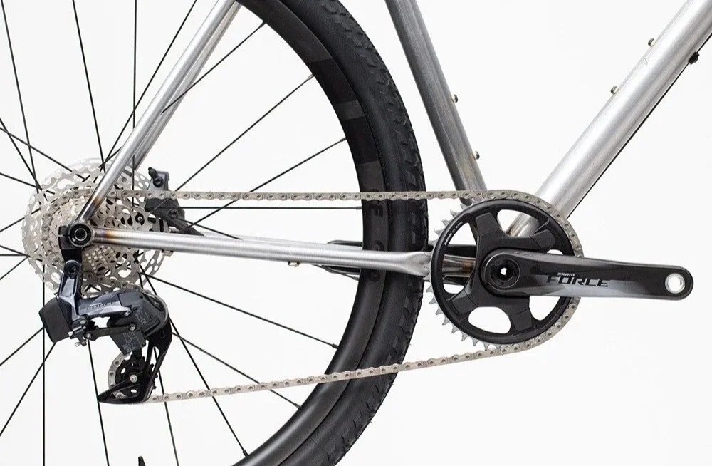 Nahaufnahme eines Fahrrads mit Fokus auf den Antrieb und das Hinterrad, silberner Rahmen, schwarzes Hinterreifen, Kettenblätter, Kette und Schalthebel.