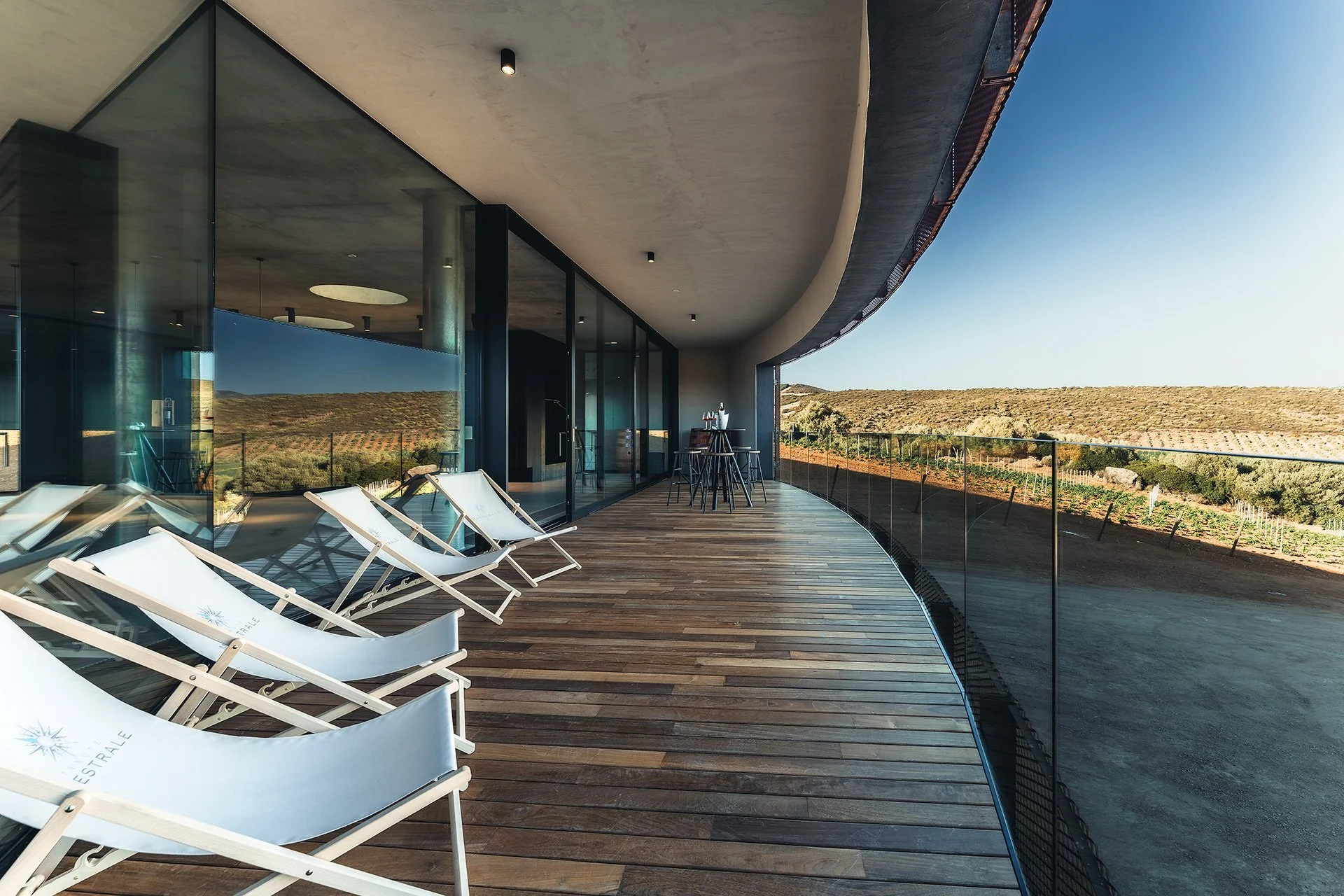 Balcone di un edificio moderno con sedie a sdraio bianche e tavoli con vista su un paesaggio rurale con colline e campi.