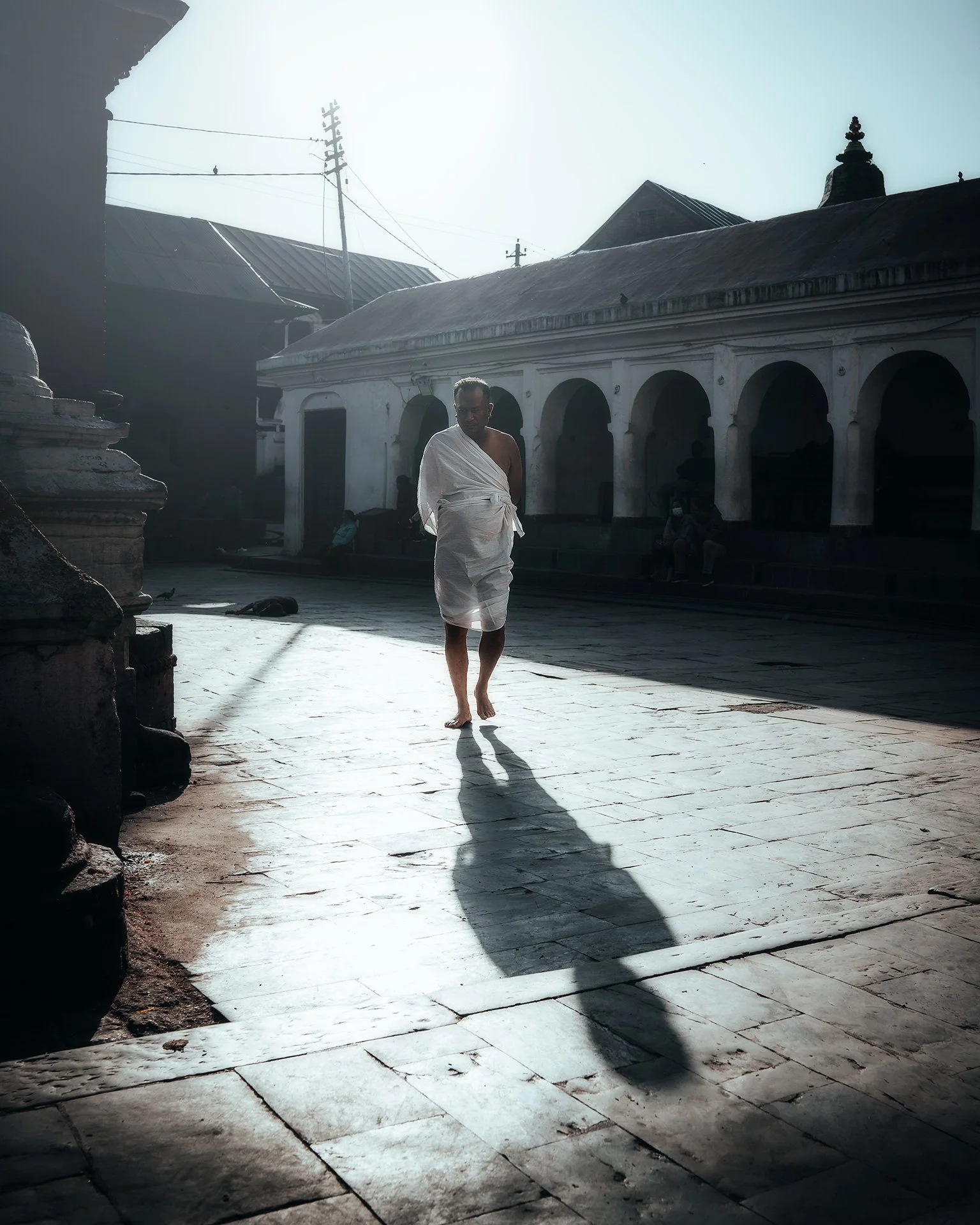Un uomo avvolto in un saree bianco cammina con passo tranquillo su un cortile di pietra, mentre il sole splende dietro un edificio storico con archi e tetto di lamiera, creando un'ombra lunga dietro di lui.