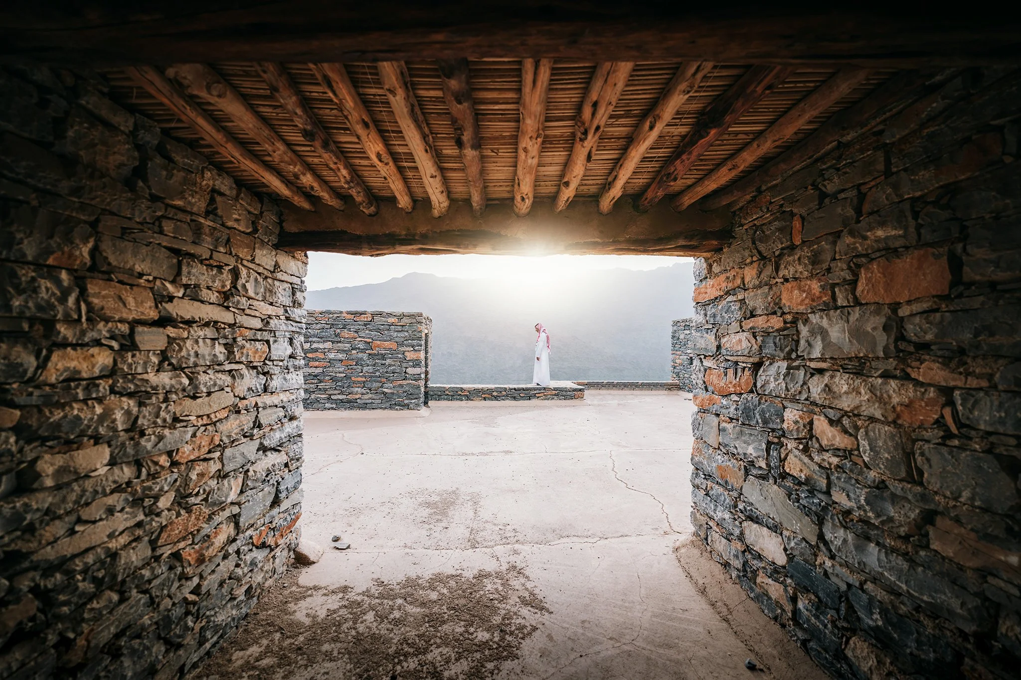 Vista attraverso un'apertura in una struttura in pietra con un soffitto in legno, con una donna in abito bianco davanti a un paesaggio montano al tramonto.