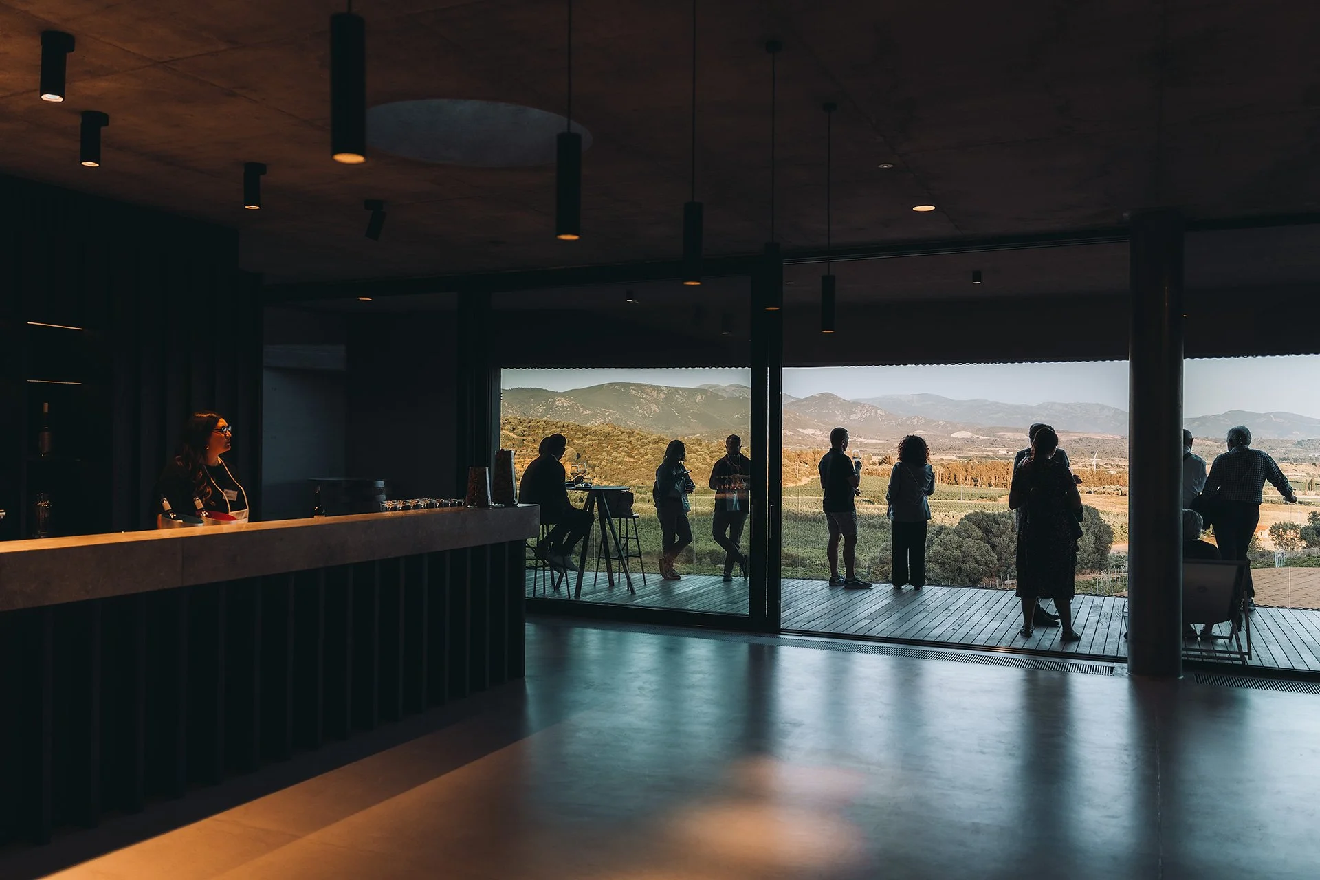 Interno di un edificio con vista panoramica su un paesaggio collinare, persone in piedi e sedute vicino a grandi finestre.