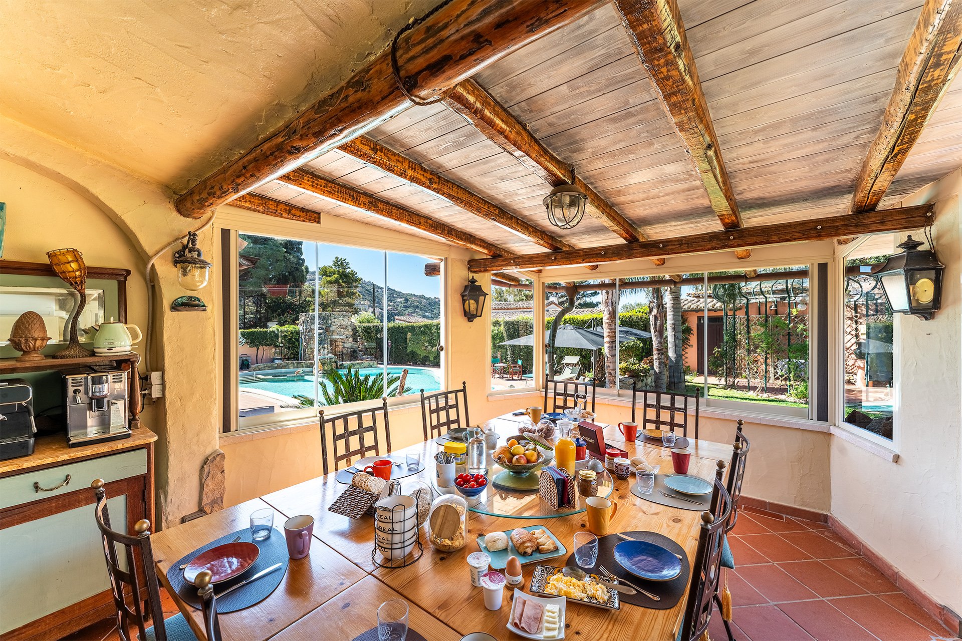 Una sala da pranzo all'interno con un tavolo apparecchiato con colazioni e bevande, grande finestra che mostra un cortile con piscina, alberi e ombrelloni.