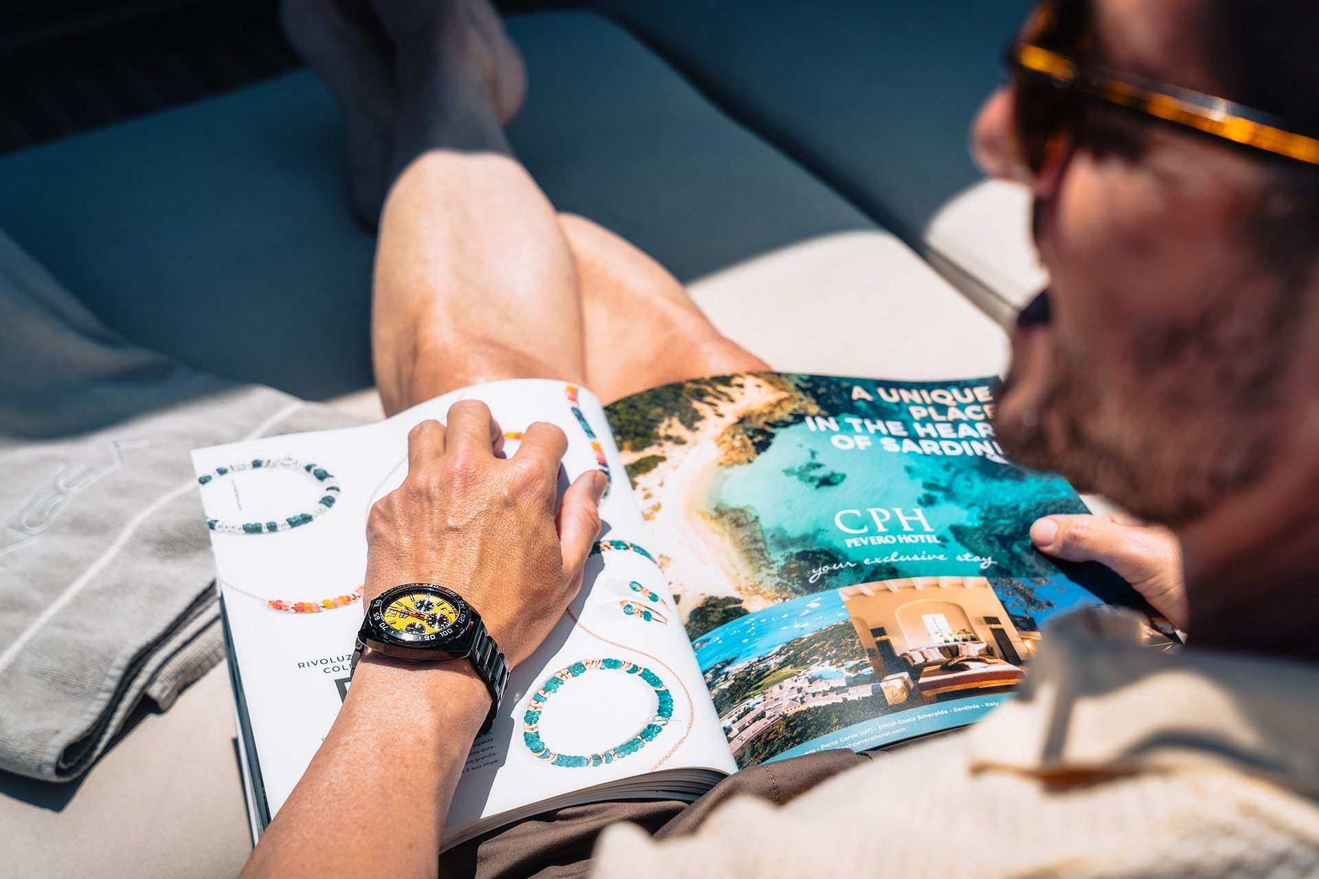 Persona che sfoglia una rivista con immagini di gioielli e destinazioni turistiche di Sardegna, mentre è seduta all'aperto. Sono visibili un orologio sul polso e alcuni accessori. La scena suggerisce relax e viaggi.