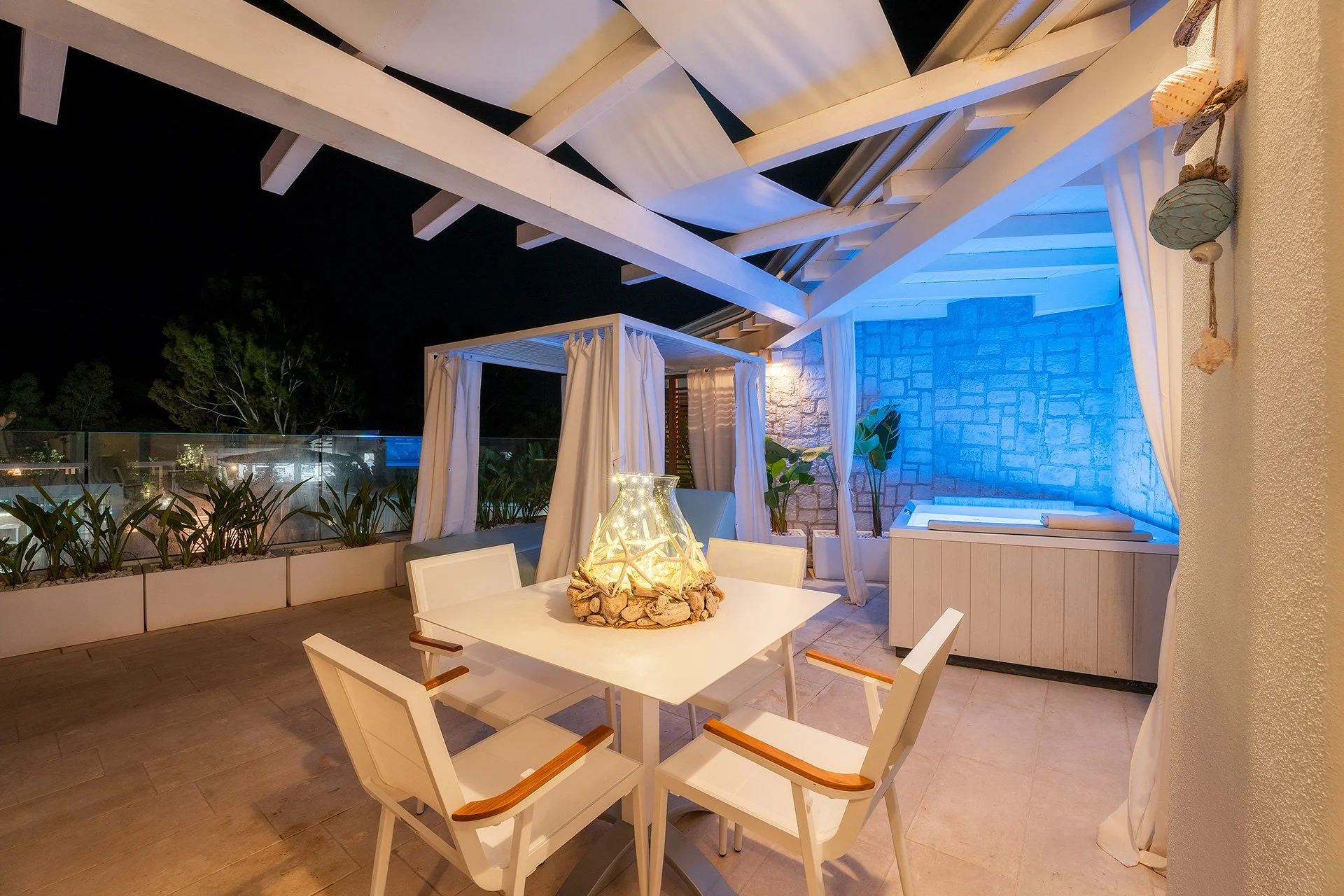 Terrazza all'aperto durante la notte con tavolo, sedie e decorazioni luminose, abbellita con tende bianche, piante verdi e luci blu su una parete di pietra, ambientazione rilassante e moderna.