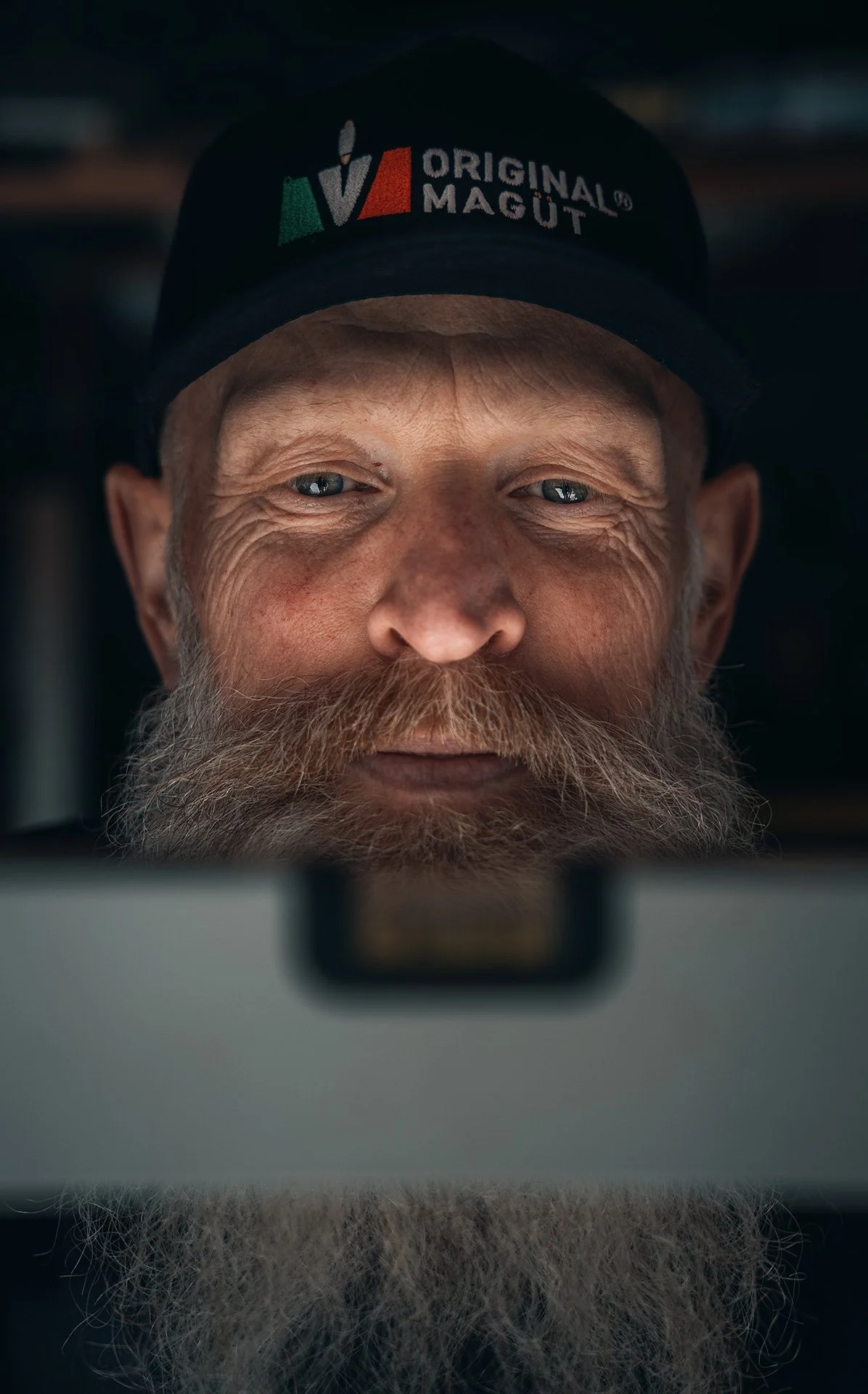 Uomo anziano con barba e cappello con logo 'Original Magüt' che guarda uno schermo