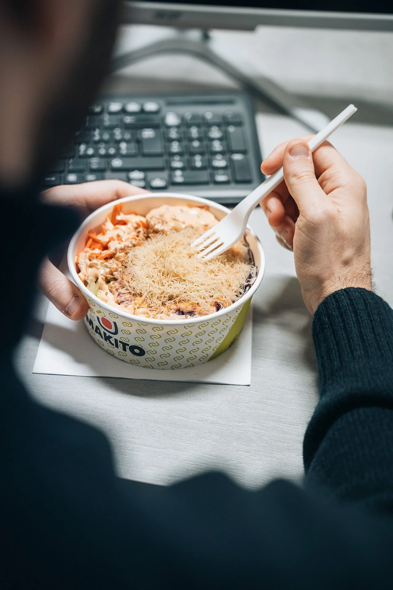 Persona che mangia un piatto di pasta con formaggio grattugiato, con una forchetta, seduto davanti a una tastiera computer