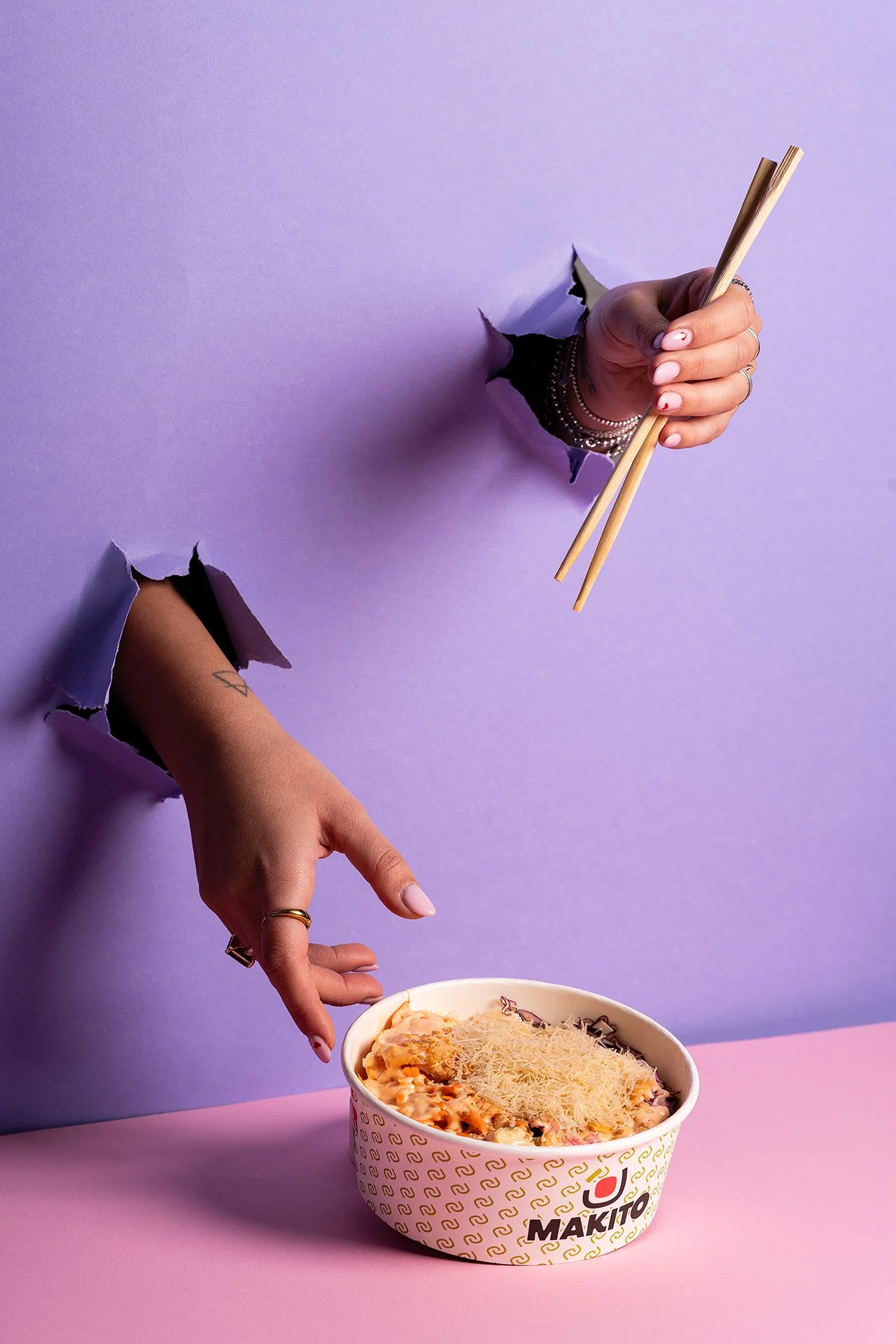 Uno strano scenario con due mani che emergono da fori in un muro viola, una mano tiene delle bacchette di legno e l'altra mano si sta avvicinando a una ciotola di cibo con formaggio grattugiato sopra, sul tavolo rosa.