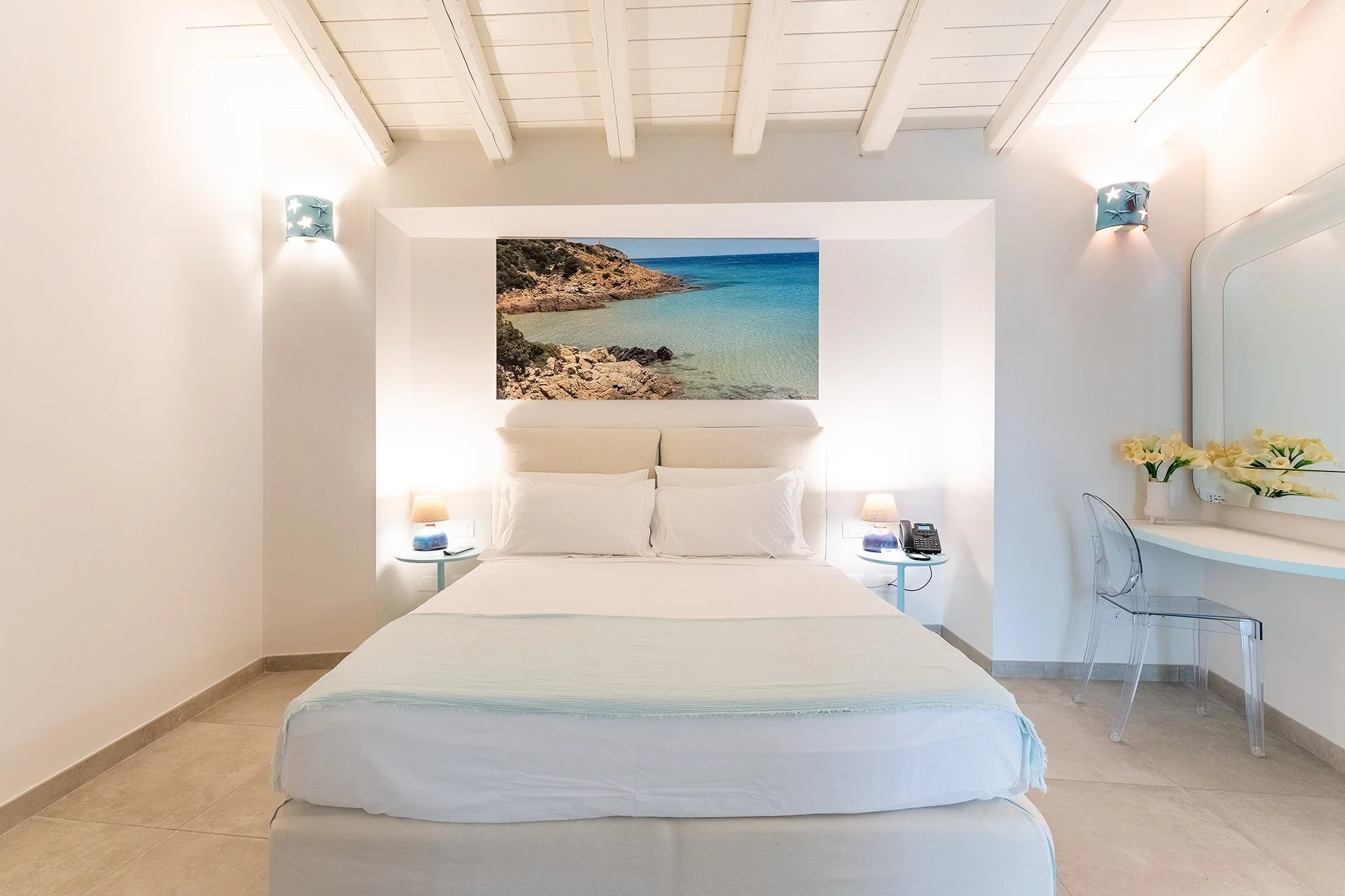 Camera da letto moderna con pareti bianche, un quadro di una spiaggia sul muro dietro il letto, due lampade ai lati, una piccola lampada sulla comodino e un telefono. Sul lato destro, una scrivania con un vaso di fiori e uno specchio.
