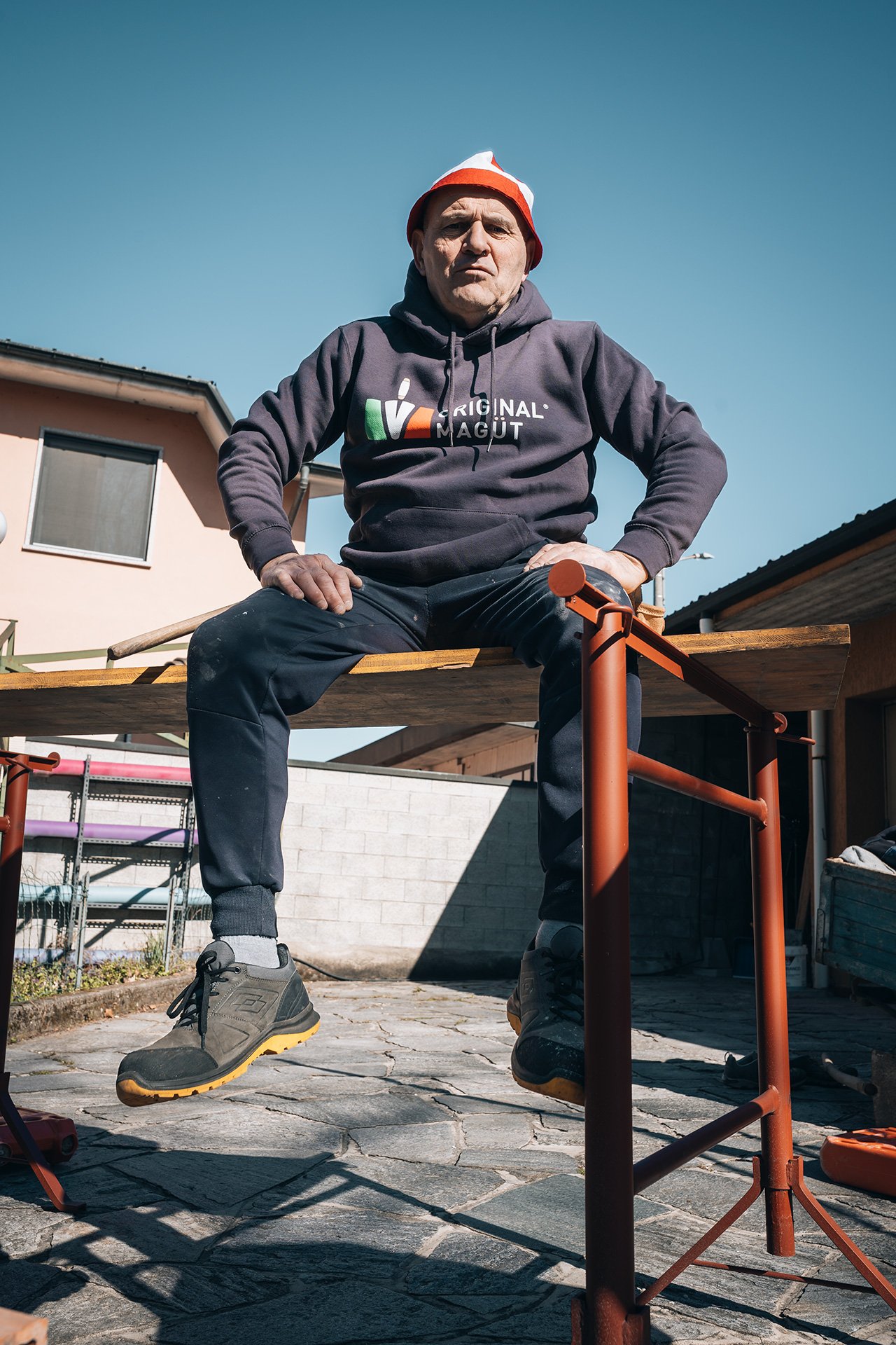 Uomo seduto su una piattaforma in legno, indossa una felpa con cappuccio grigia, pantaloni casual e scarpe da trekking, con un cappello rosso e bianco, in un cortile soleggiato con case e cielo azzurro sullo sfondo.