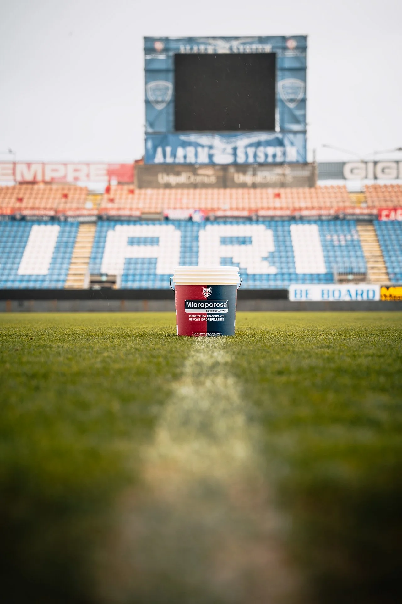 Una secchia di vernice rossa con scritte italiane e il logo del Cagliari Calcio posizionata su un prato all'interno di uno stadio vuoto, con lo sfondo di gradinate e un grande schermo.