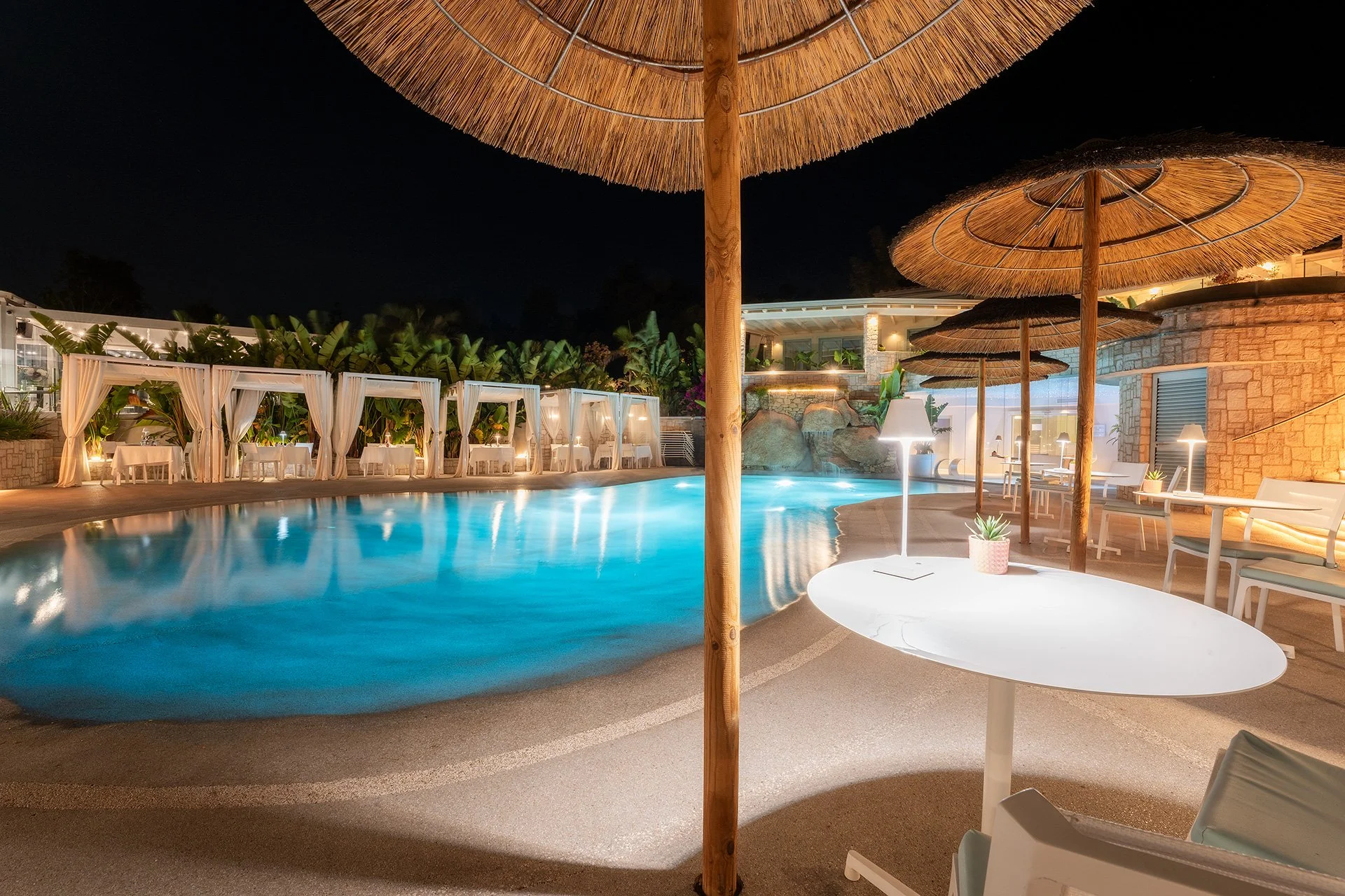 Piscina illuminata di notte con ombrelloni di paglia, sedie e lampade nelle vicinanze in un ambiente di resort o hotel.