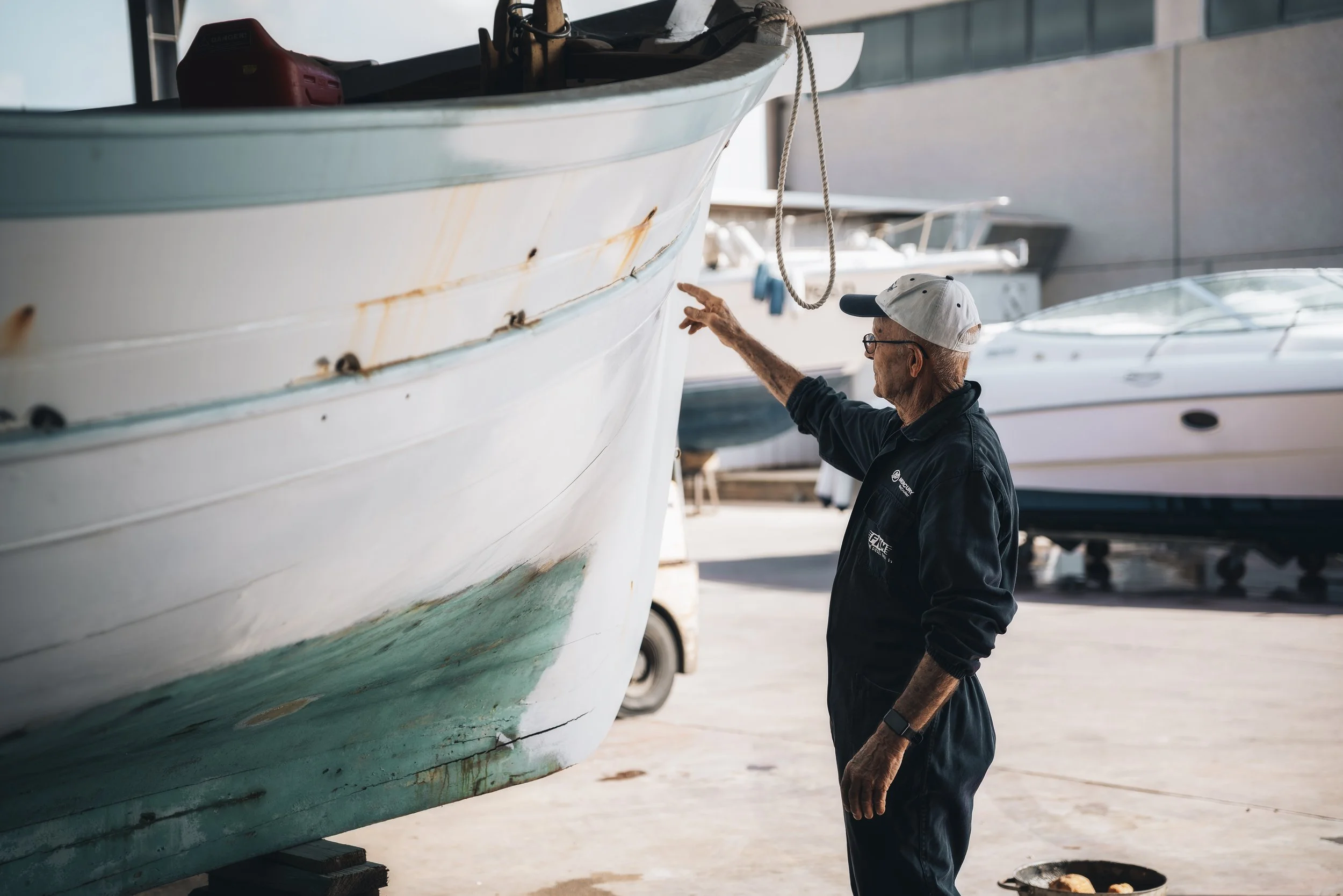 Un uomo anziano con cappellino bianco e occhiali sta ispezionando una barca bianca in un capannone di ormeggio, in un cantiere navale, con altri yacht sullo sfondo.