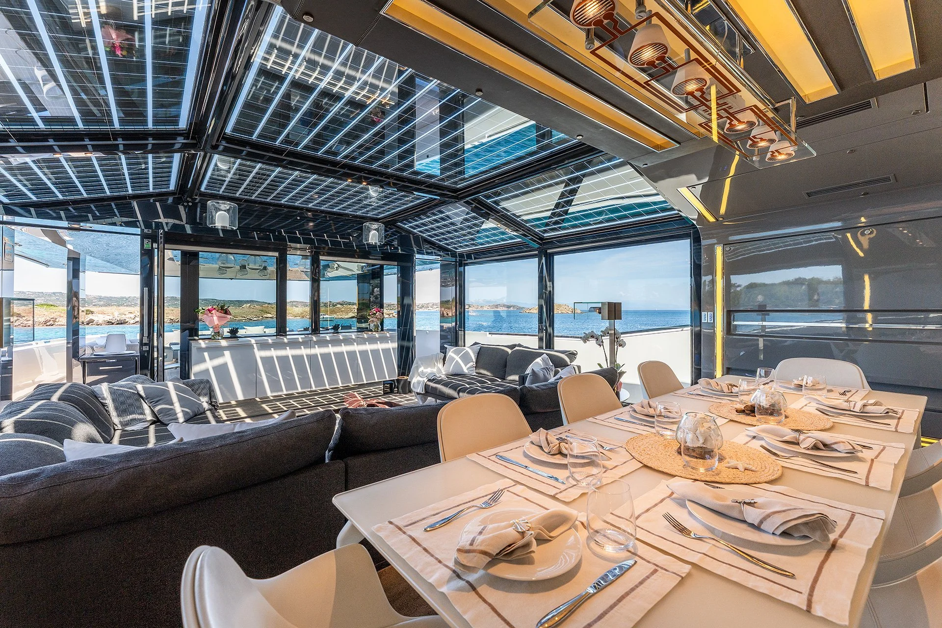 Interno di uno yacht con vista sul mare, arredamento moderno e accogliente, tavoli apparecchiati con piatti, bicchieri e tovaglioli, divani neri e sedie beige, grandi finestre con vista panoramica sul mare e sul paesaggio costiero.