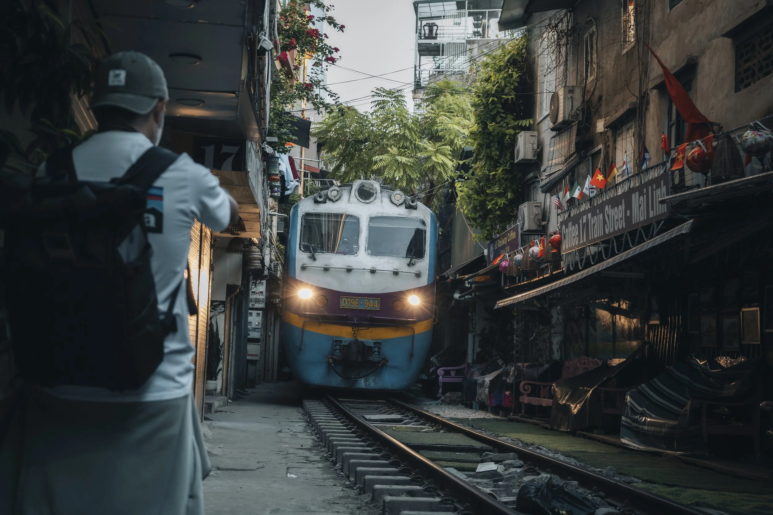 Una persona con cappello e zaino guarda un treno blu in una strada stretta circondata da edifici e negozi decorati con bandiere. Sullo sfondo ci sono alberi e un cielo chiaro.