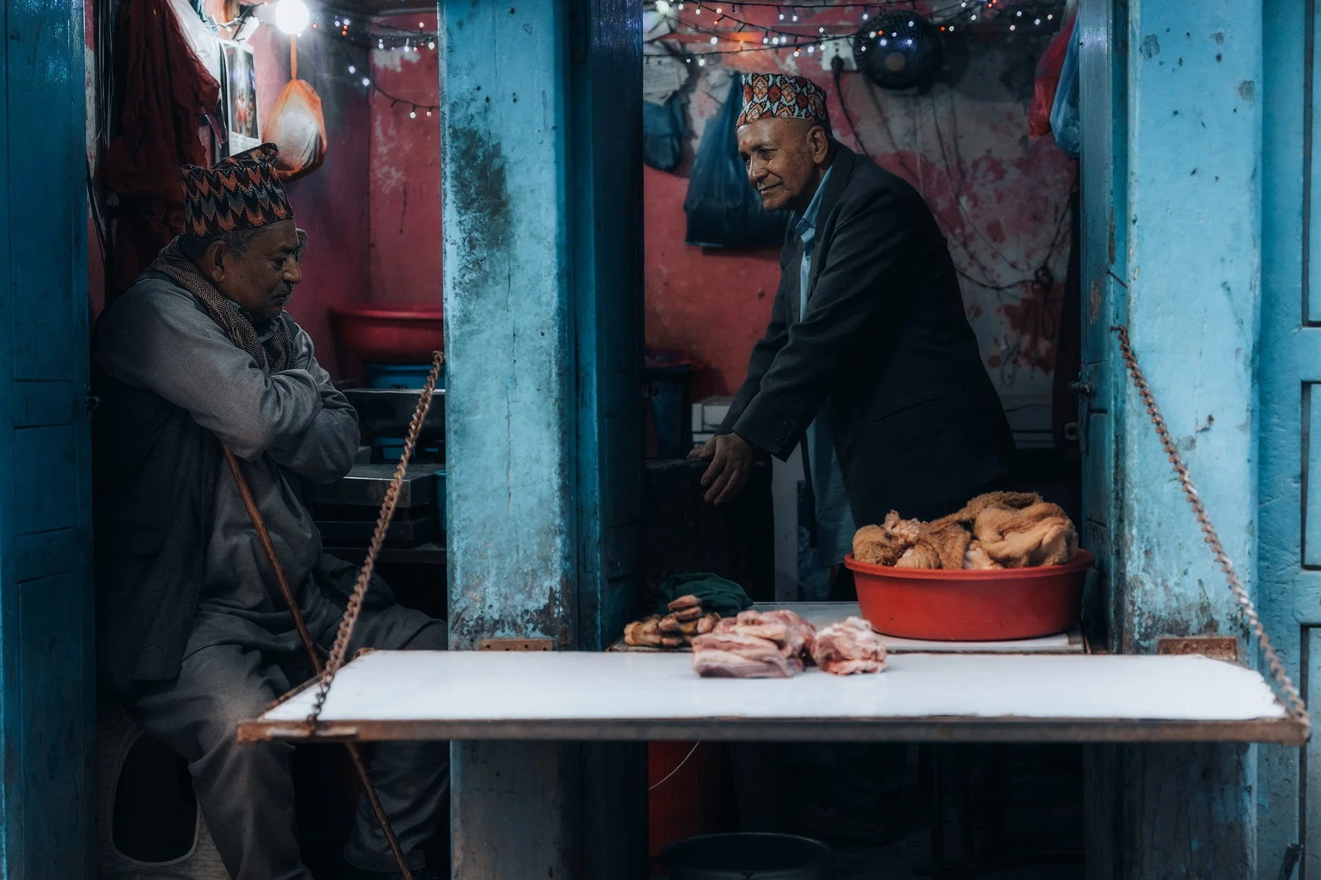 Due uomini con cappelli tradizionali nepalesi vendono carne in un mercato al coperto. Uno è seduto con le braccia incrociate, l'altro si inclina verso un bancone con pezzi di carne su un tavolo e una grande ciotola di carne davanti a lui. Le pareti s