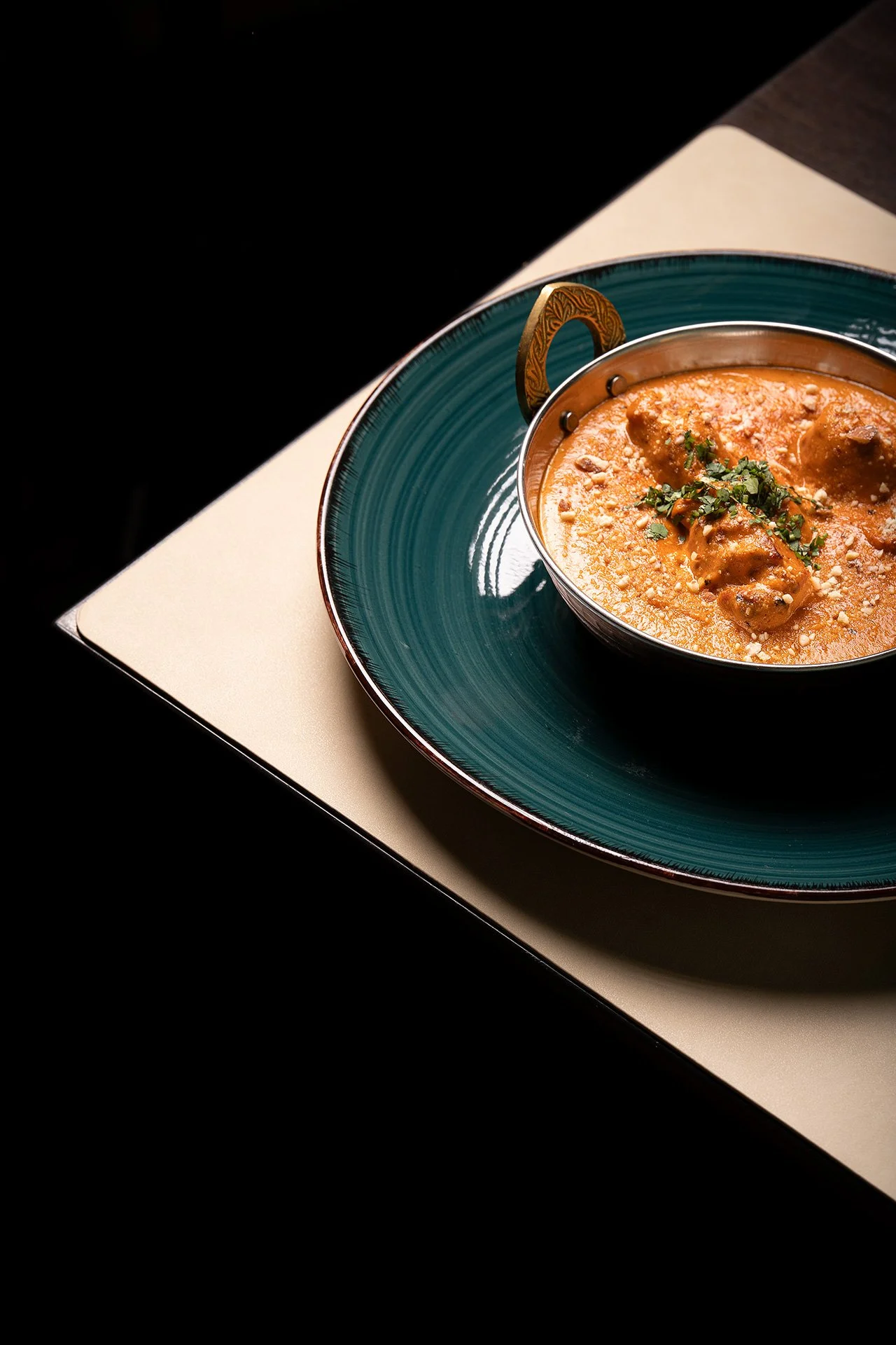Una ciotola di zuppa calda con carne e verdure, decorata con erbe fresche, su piatto di colore verde/blu scuro, posato su un vassoio bianco e sfondo nero.