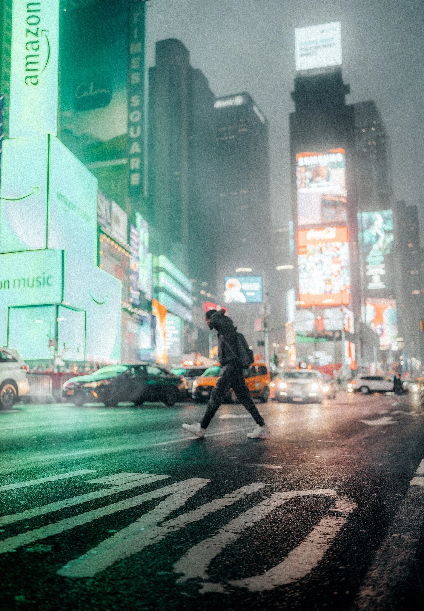 Una persona attraversa la strada a Times Square di notte, con luci al neon e grattacieli illuminati sullo sfondo, sotto la pioggia.