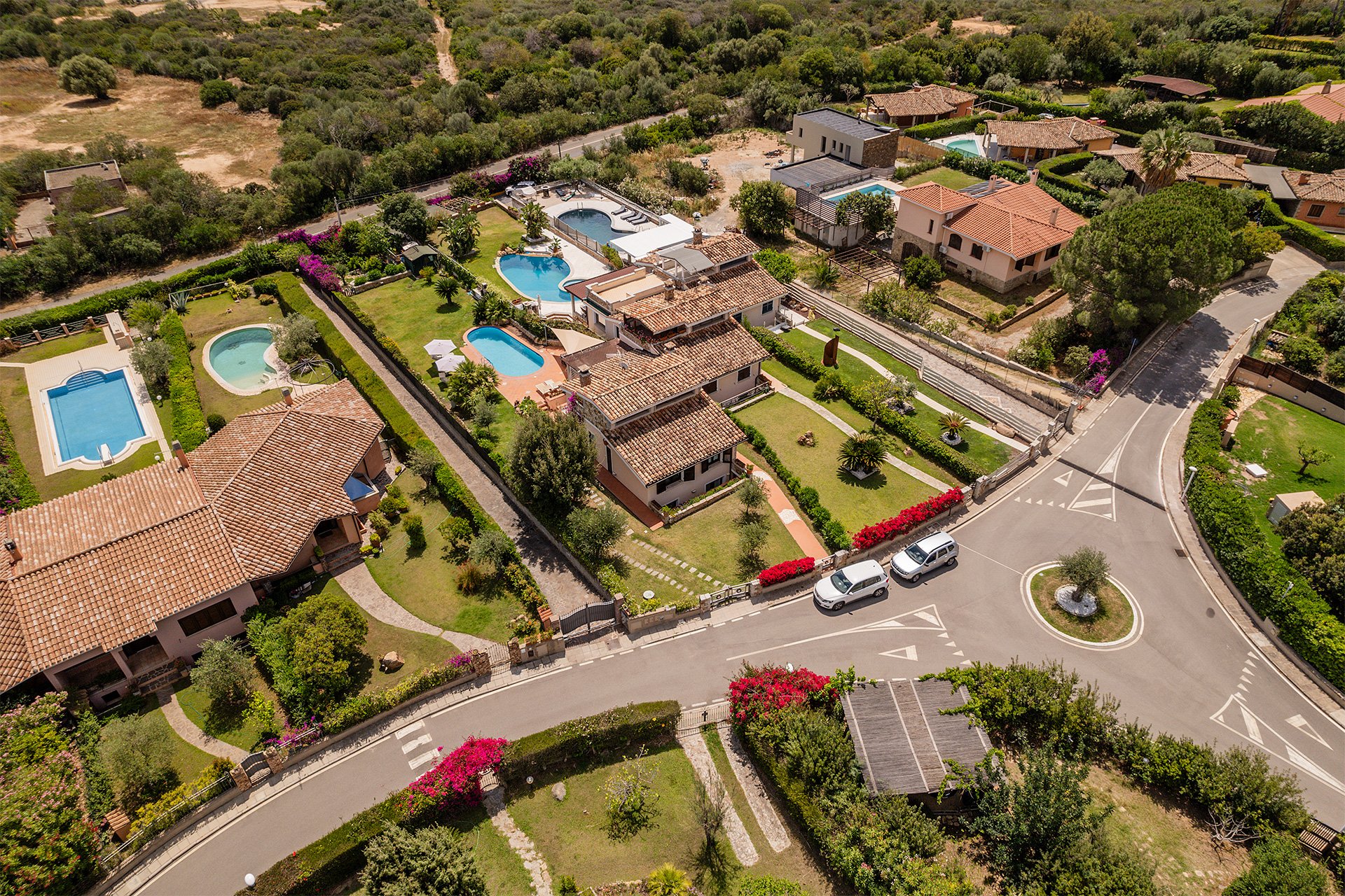 Vista aerea di una zona residenziale con case, piscine, giardini ben curati, alberi e un'area naturale alle spalle. Una strada curva passa davanti alle case, con alcuni veicoli parcheggiati e un'area di rotonda con un albero centrale.