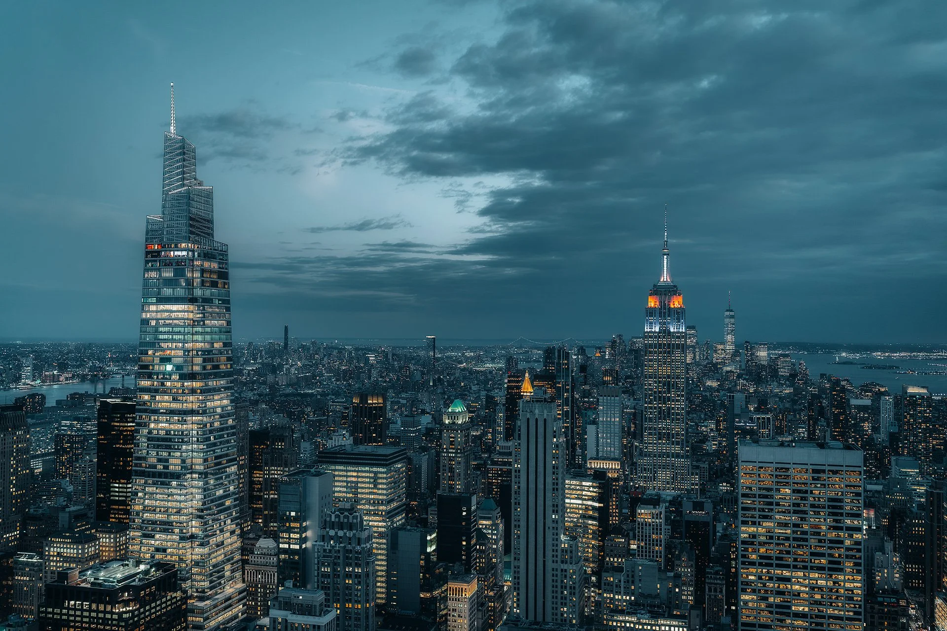 Vista notturna dello skyline di New York con grattacieli illuminati, tra cui l'Empire State Building, sotto un cielo nuvoloso.