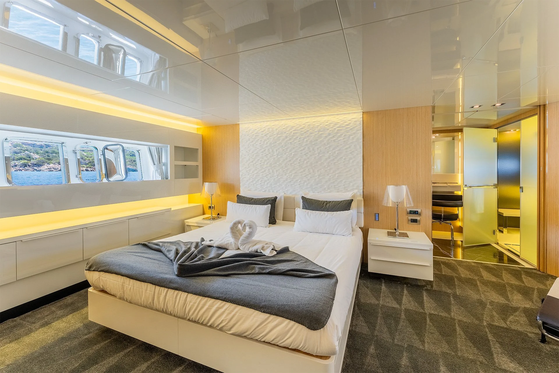 Camera da letto moderna con finestra vista mare, letti con asciugamani a forma di oche, arredamento in legno chiaro, parete decorativa bianca, lampade ai lati del letto, bagno in dettaglio, colori neutri con accenti gialli.