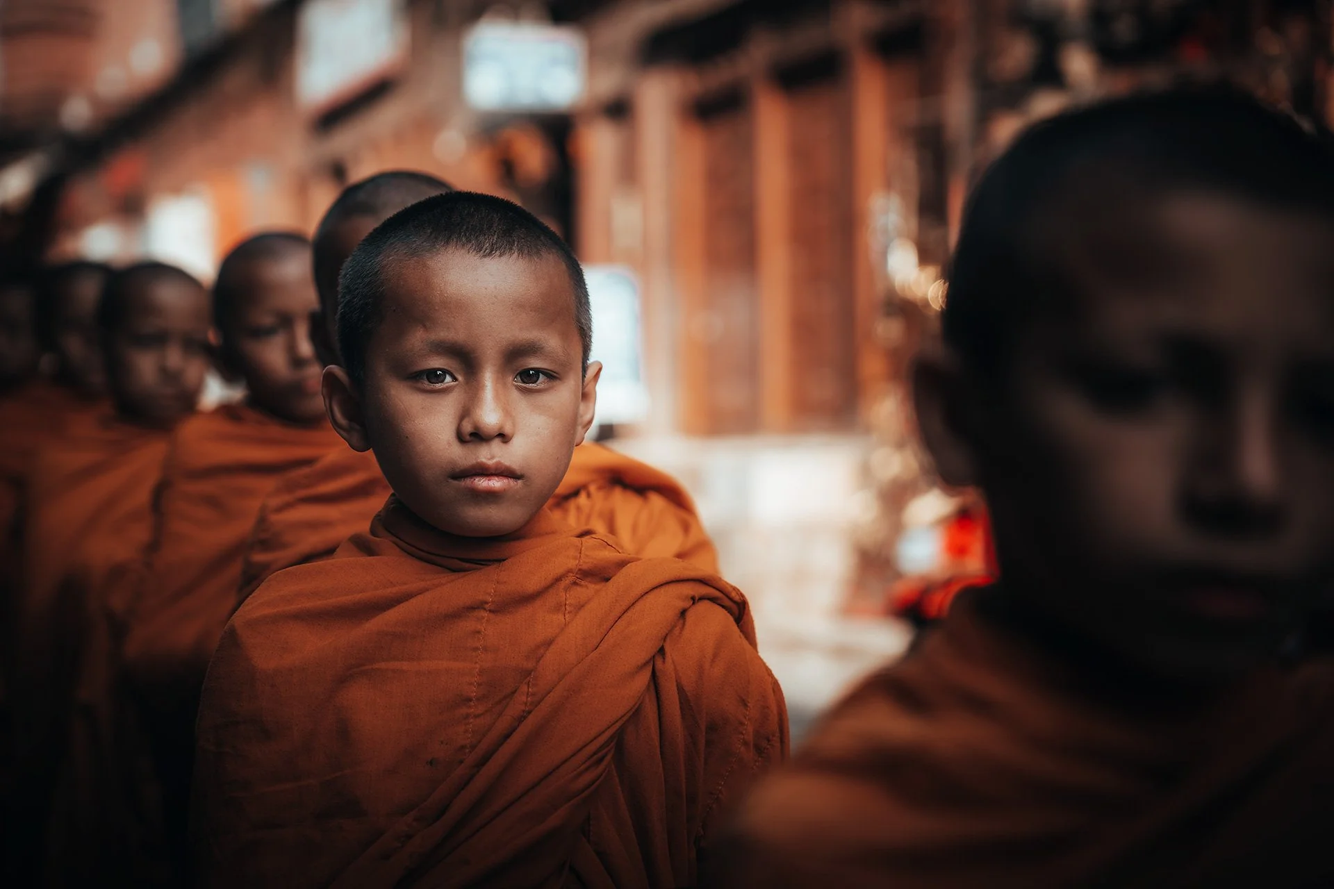 Ragazzini monaci buddhisti con sandali arancioni seduti in fila in un'area interna di un tempio, viso del primo ragazzino in primo piano, sfondo sfocato con altri monaci.