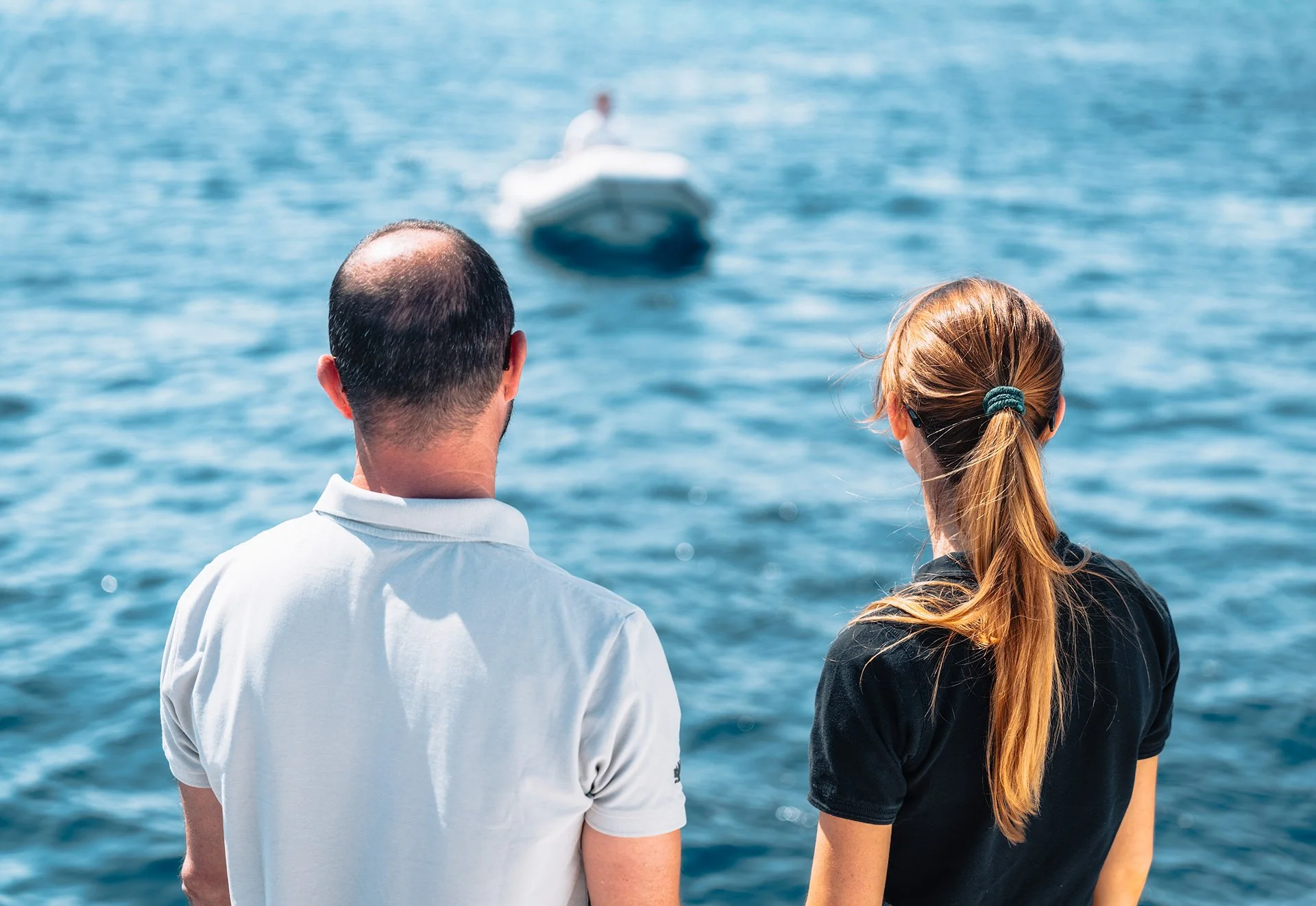 Due persone, un uomo e una donna, osservano un mare o un lago mentre si trovano sulla riva. Sullo sfondo, una barca a motore in acqua.