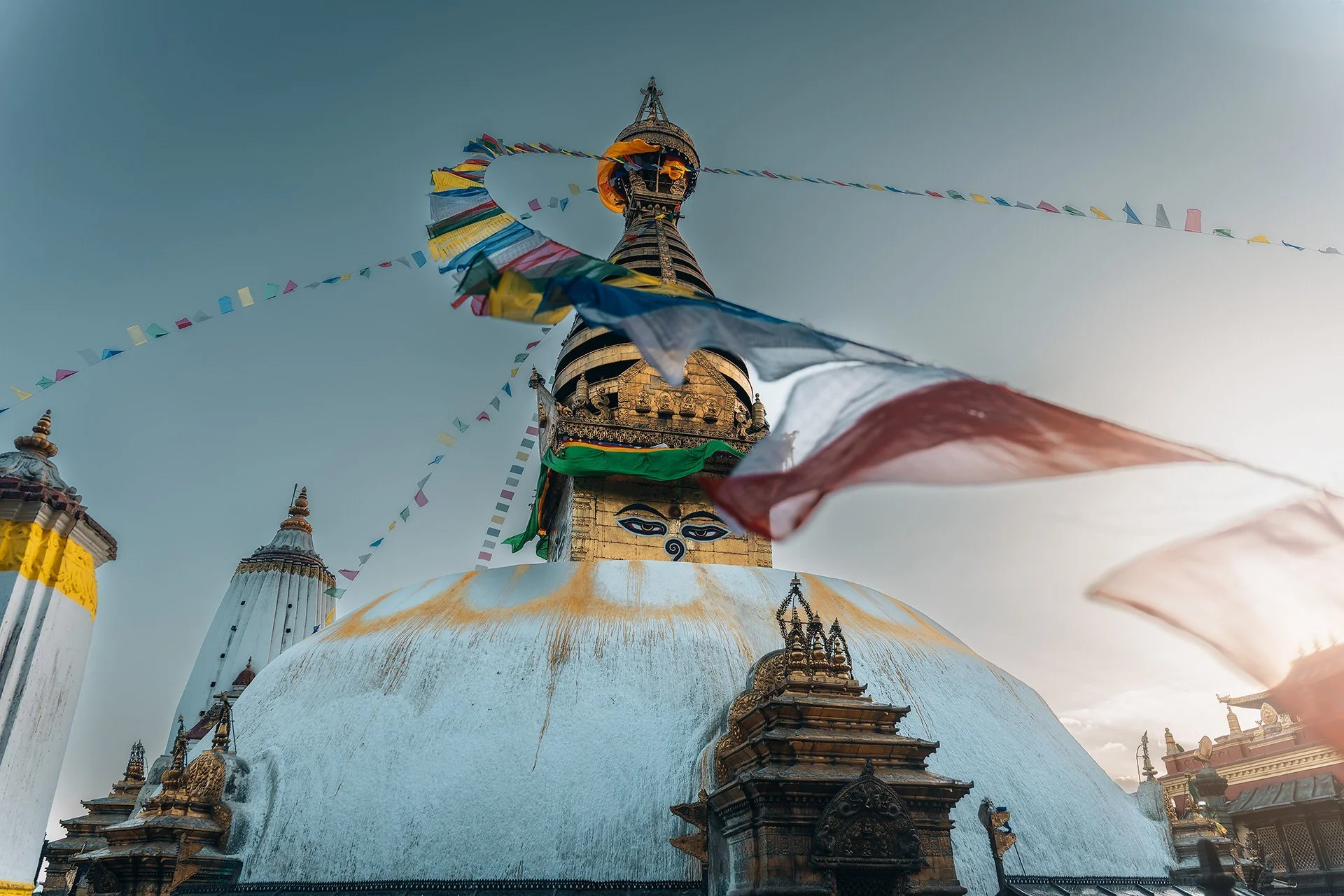 Vista della stupa di Swayambhunath con bandiere di preghiera che svolazzano al vento e un cielo chiaro sullo sfondo, in Nepal.