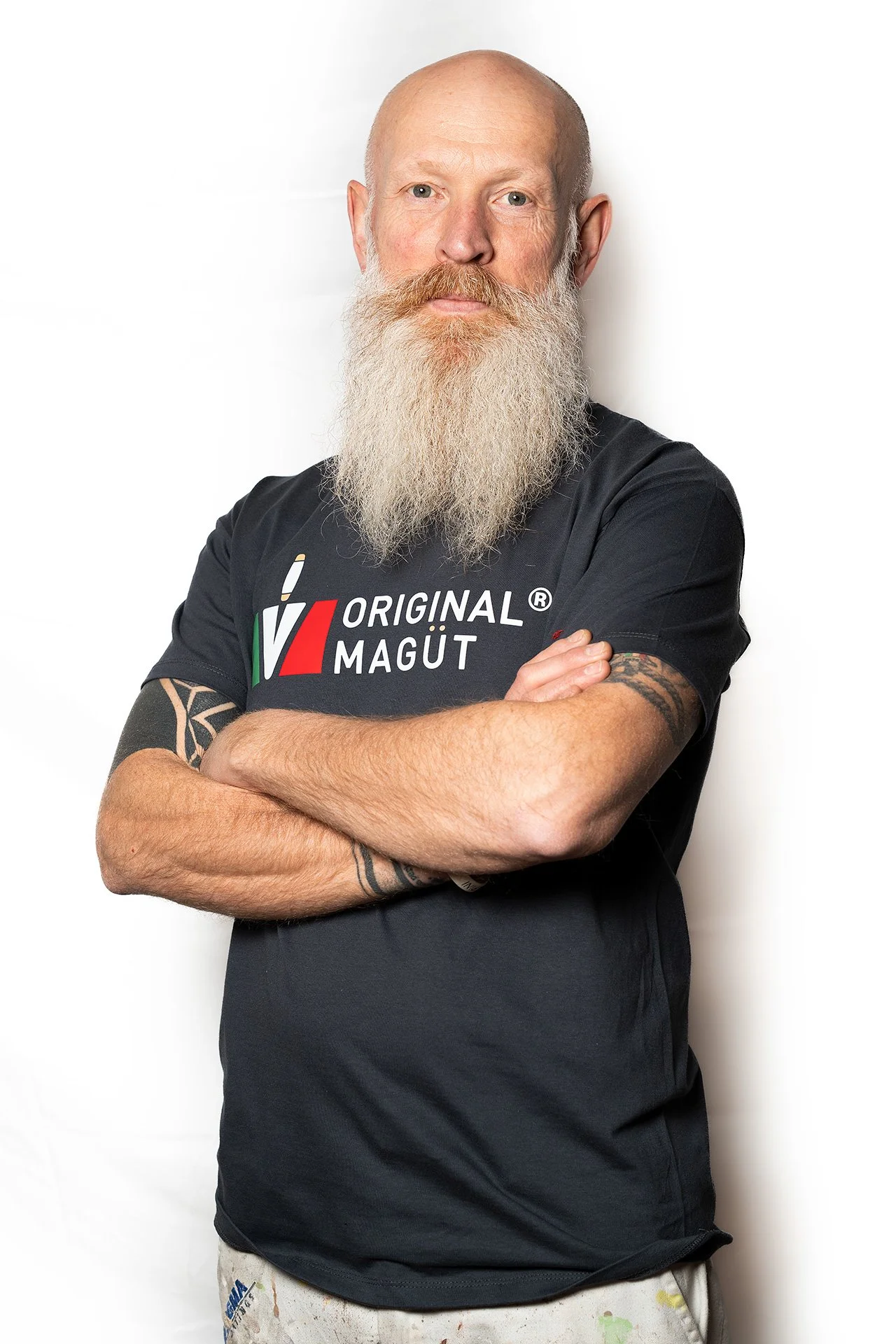 Uomo con barba lunga e tatuaggi, vestito con maglietta nera con scritto "Original Magut", braccia incrociate, sfondo bianco.