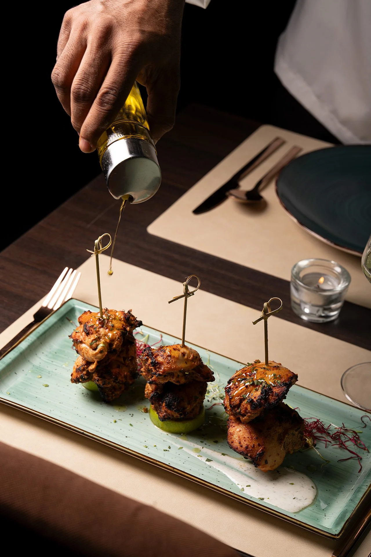 Mano che versa olio su spiedini di carne alla griglia, serviti su un piatto. Tavola appare elegante con posate, bicchiere e candele.