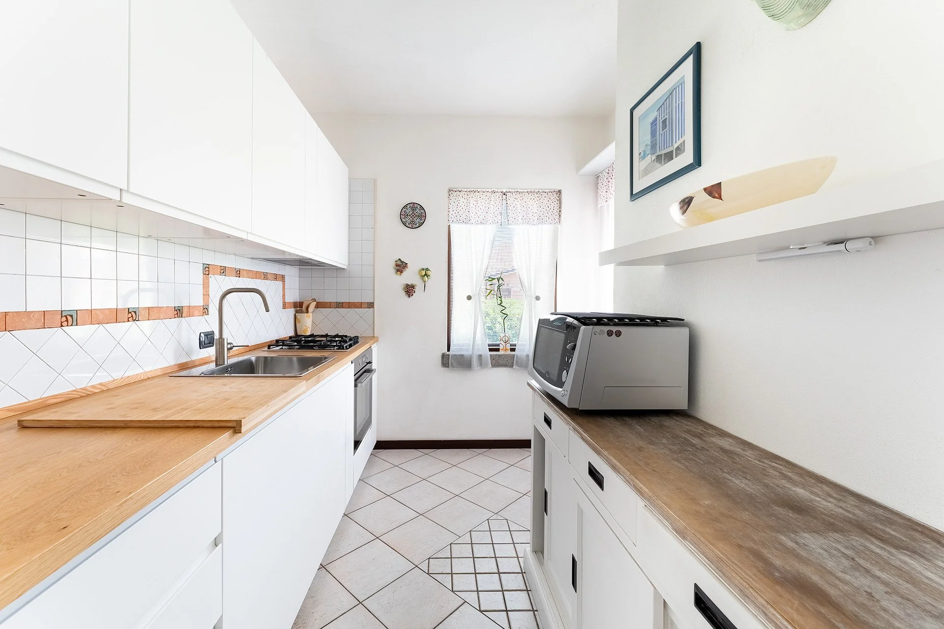 Cucina moderna con piano di lavoro in legno, fornelli a gas, forno, lavabo e pareti bianche decorate con piastrelle e quadri. Finestra con tende a fiore. Stereo appoggiato su un mobile.