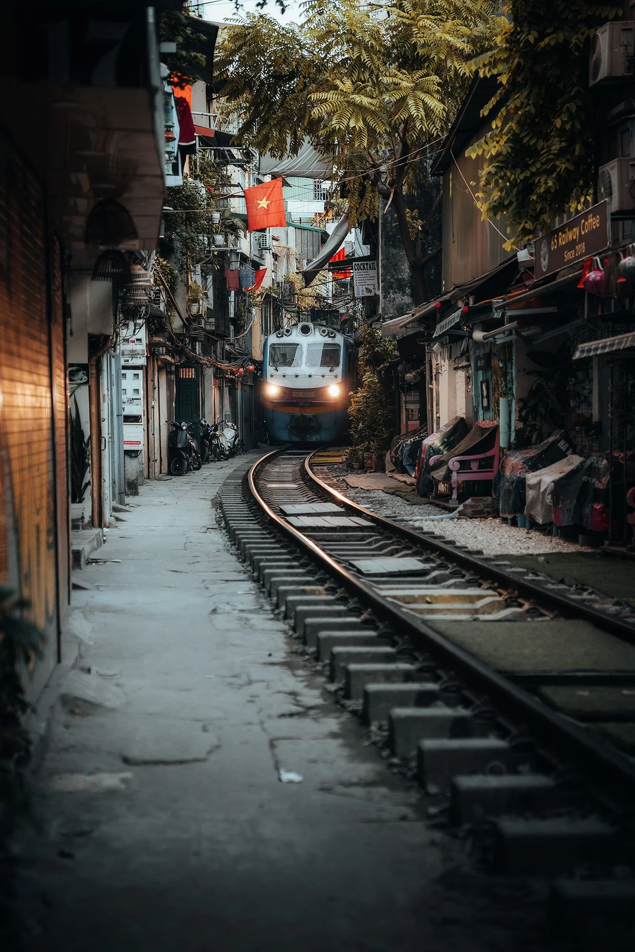 Treno che passa tra case e negozi in una stradina stretta, con arcobaleni di luci e bandiere vietnamite.