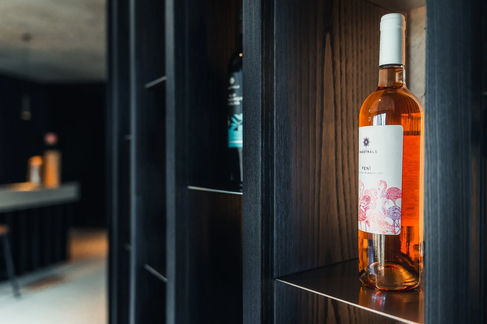 Bottiglia di vino rosa con etichetta decorativa di fenicotteri, posizionata su una mensola di legno scuro in uno spazio ambientale moderno.
