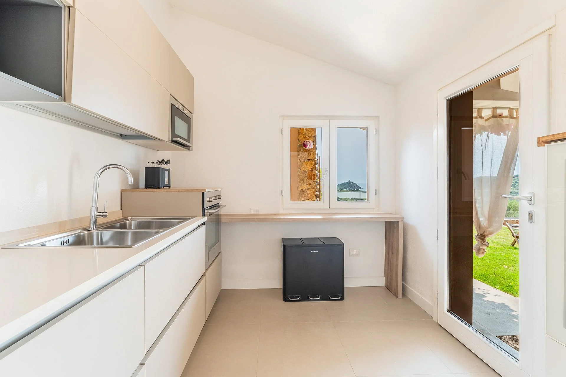 Cucina moderna con lavello, microonde, sedia, finestra e porta finestra che mostra un giardino verde con una sedia e un tendone in tessuto, vista sulla collina in lontananza.