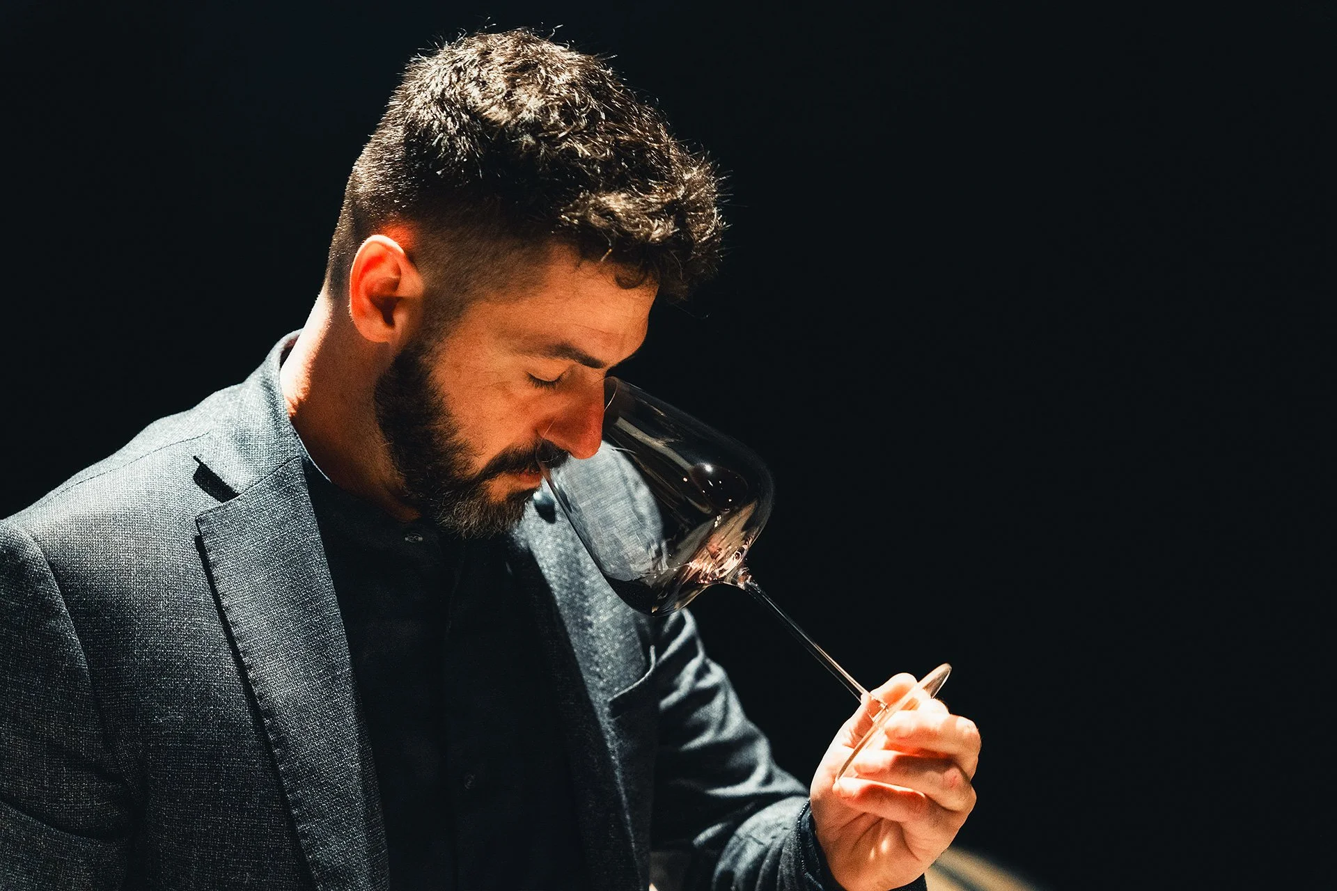 Uomo con barba e capelli ricci che sorseggia un bicchiere di vino rosso, indossa un abito grigio, sfondo nero.