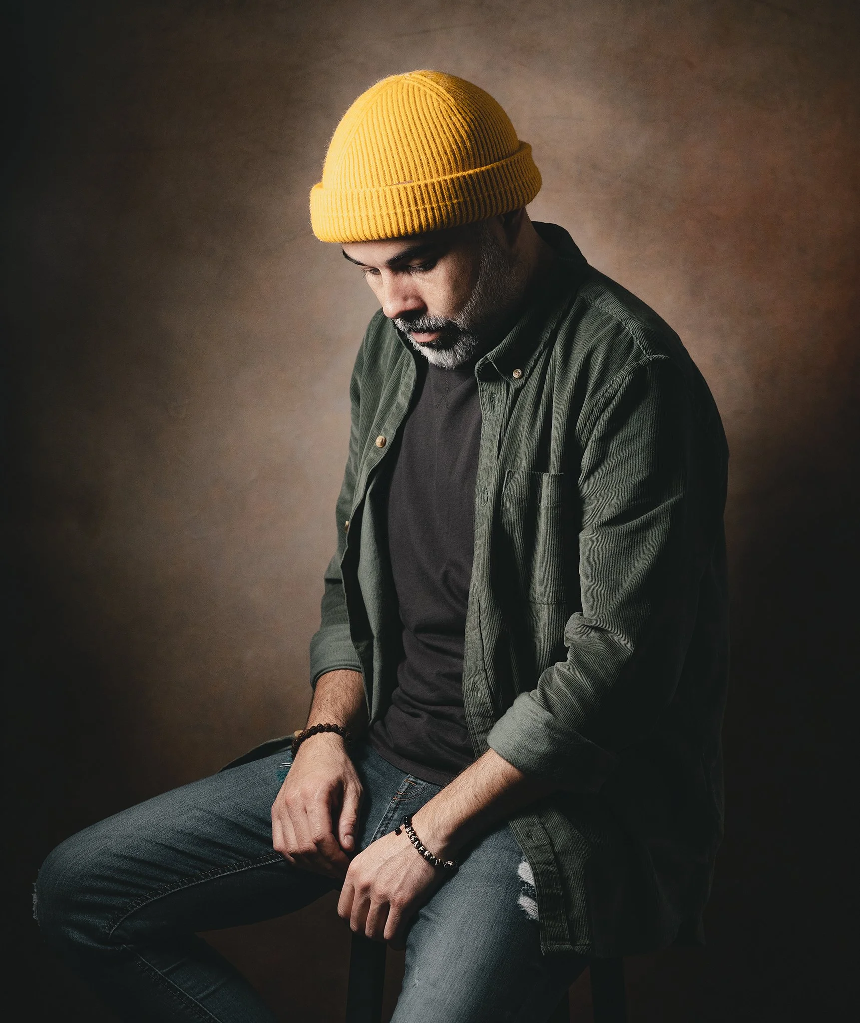 Un uomo con barba e baffi indossa un berretto giallo, una maglietta nera e una camicia verde scura, seduto su una sedia contro uno sfondo scuro e neutro, guarda verso il basso.