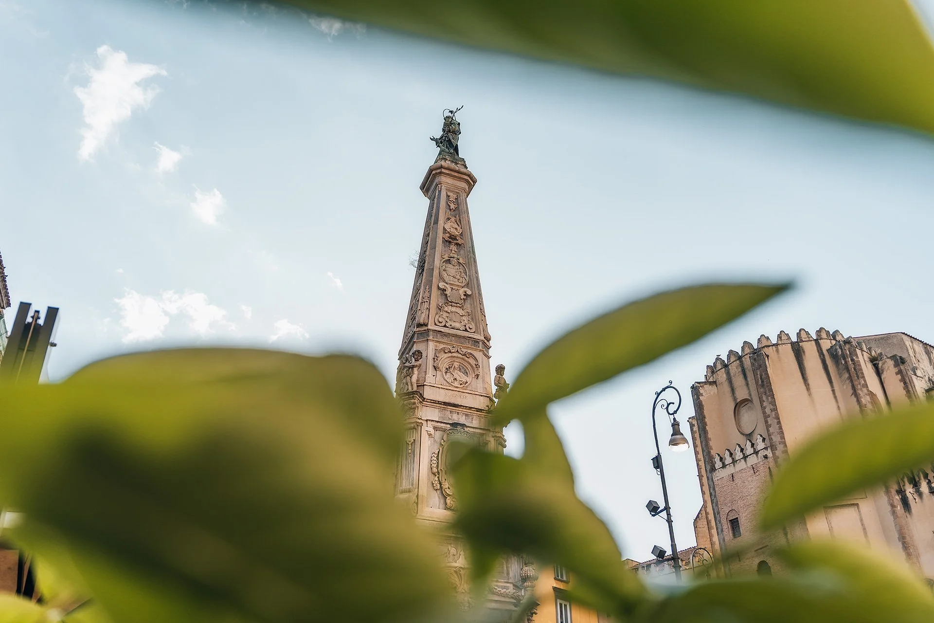 Particolare di una torre con una statua in cima, incorniciata da foglie verdi in primo piano.