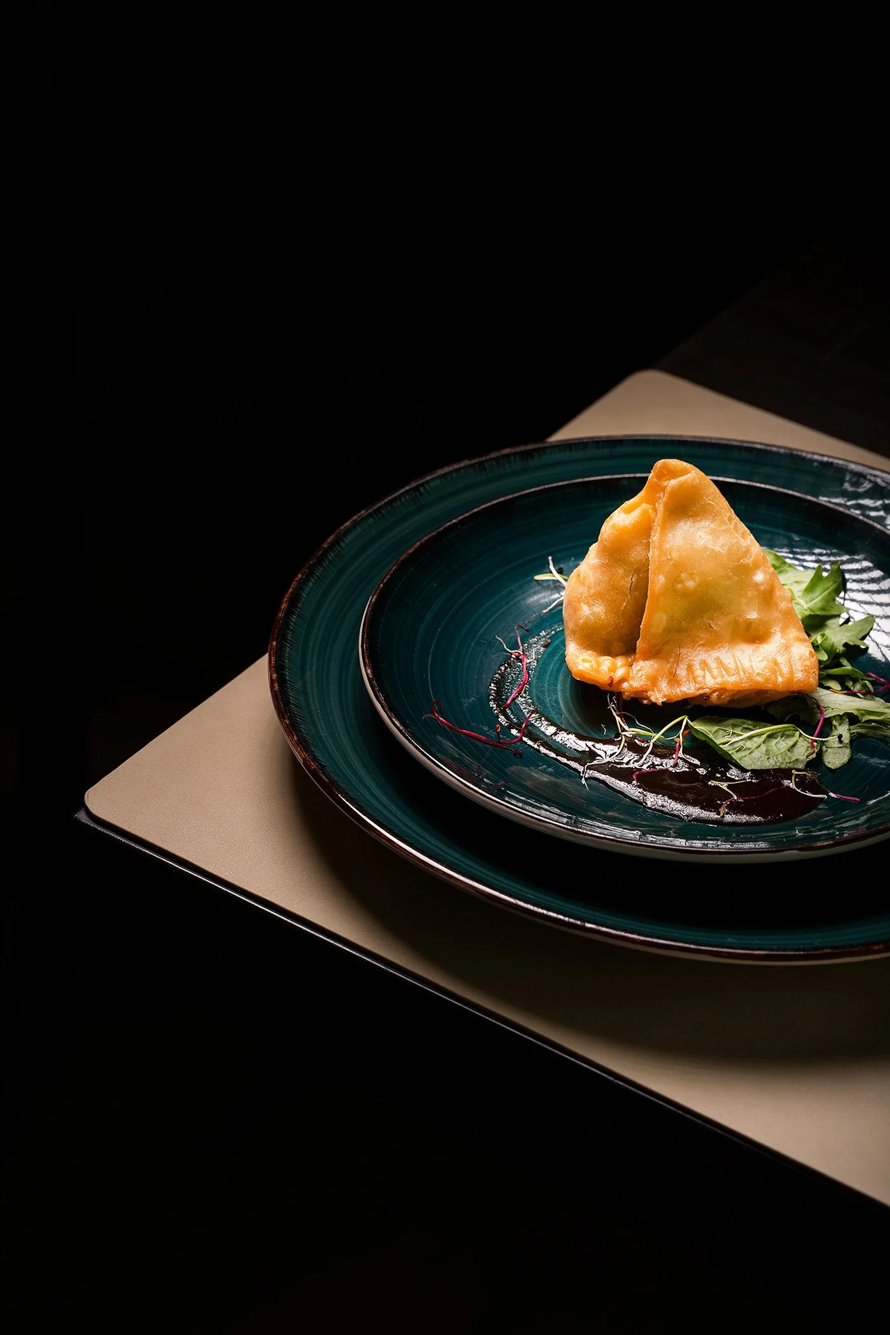 Due samosas dorate su un piatto blu con foglie di insalata e germi, sfondo nero.