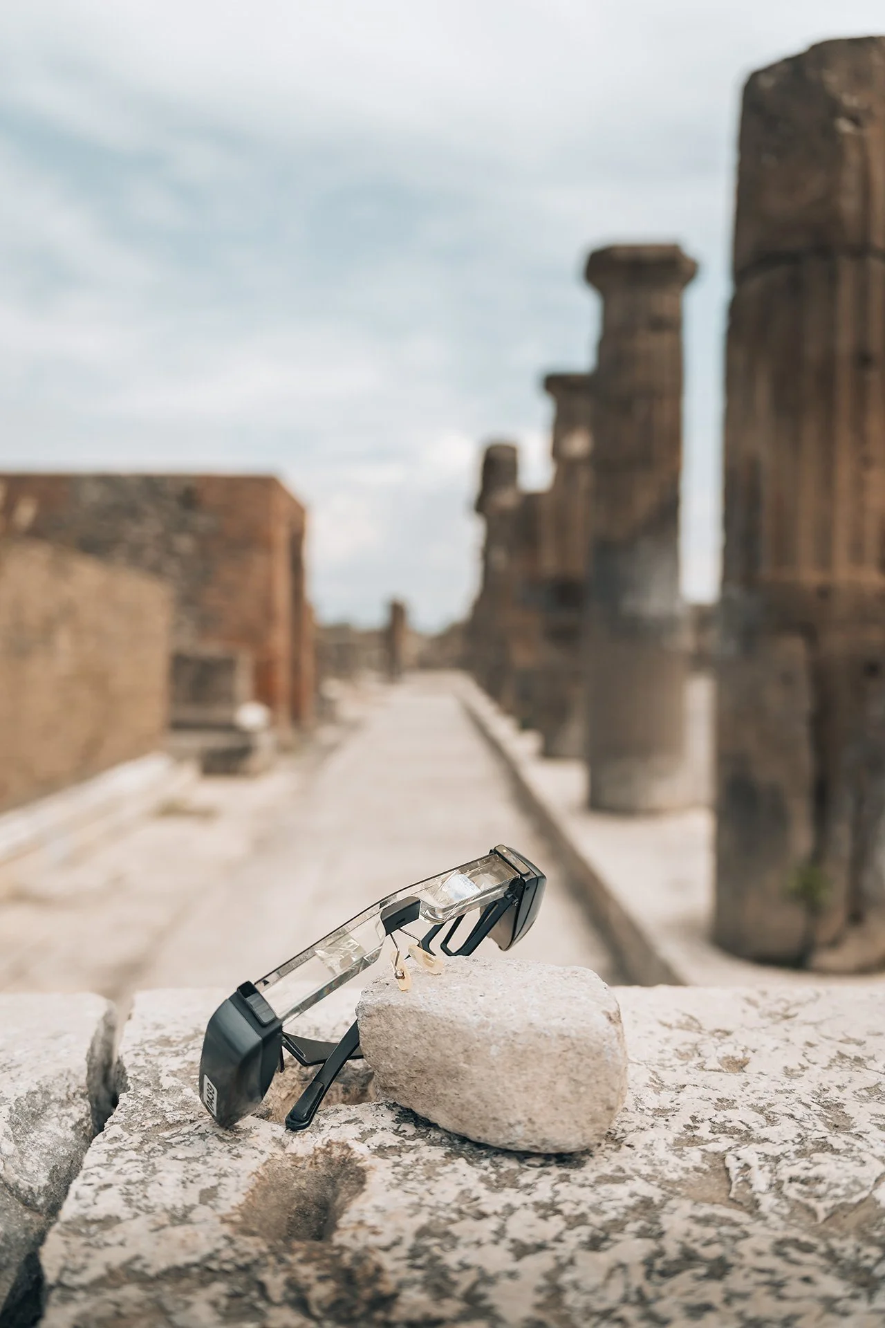 Occhiali da sole appoggiati su una pietra, con rovine antiche in sfondo sotto un cielo nuvoloso.