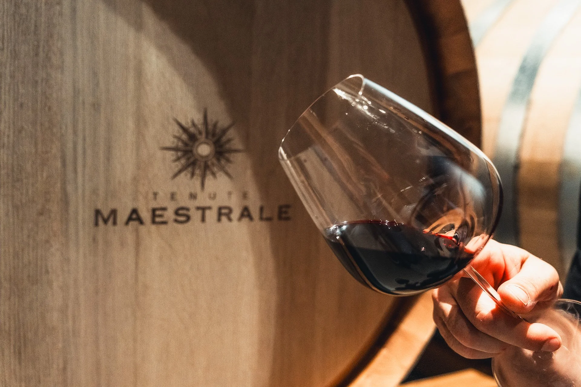 Calice di vino rosso tenuto da una mano, sfondo con il logo 'Tenuta Maestrale' su una parete di legno.