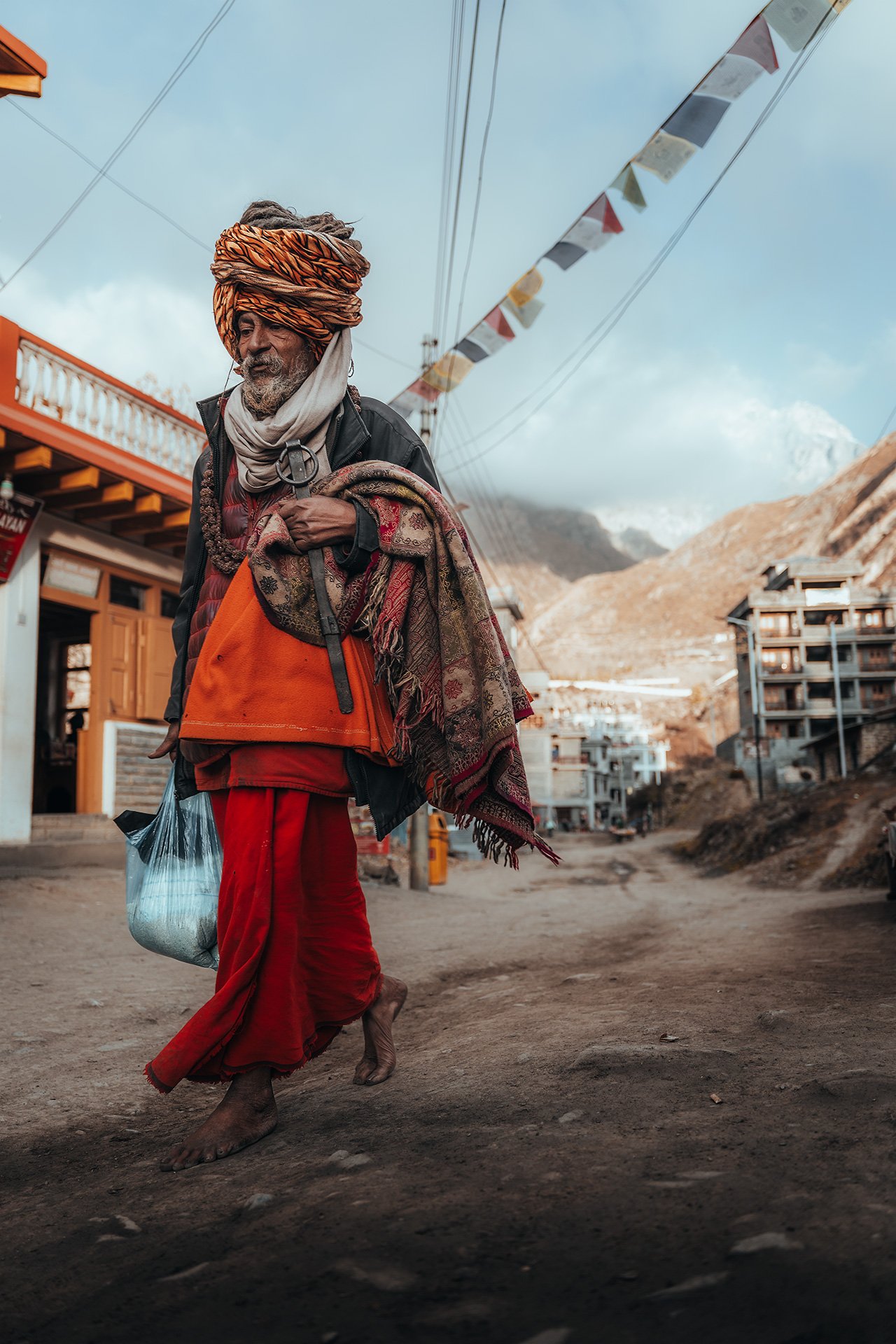 Un uomo anziano con il turbante arancione, vestito con un abito rosso e una giacca nera, cammina scalzo per le strade di un villaggio di montagna. Sta portando una borsa di plastica con quello che sembra riso e porta un drappo colorato sulle spalle. 