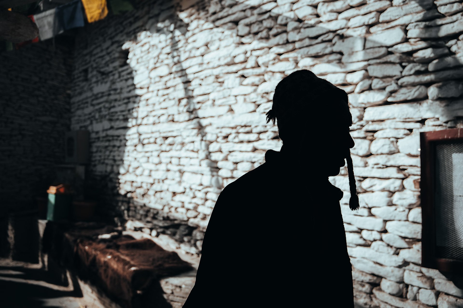 Profilo di una persona con capelli legati e barba, silhouette nera sul muro di pietra chiara, con ombra di luce solare intensa.