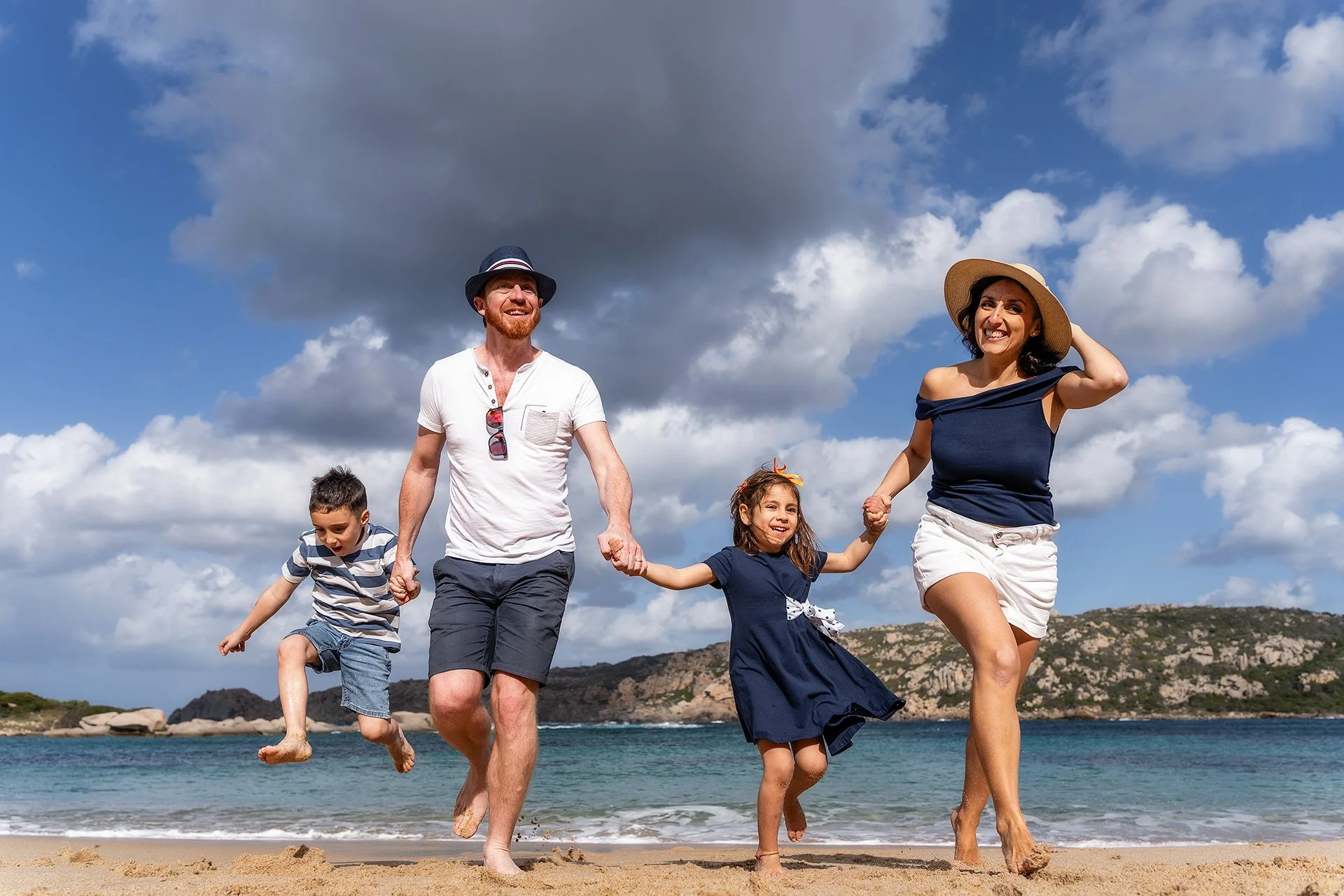 Una famiglia di quattro persone che cammina sulla spiaggia, sorridendo e tenendosi per mano, con il mare e le montagne sullo sfondo, sotto un cielo nuvoloso.