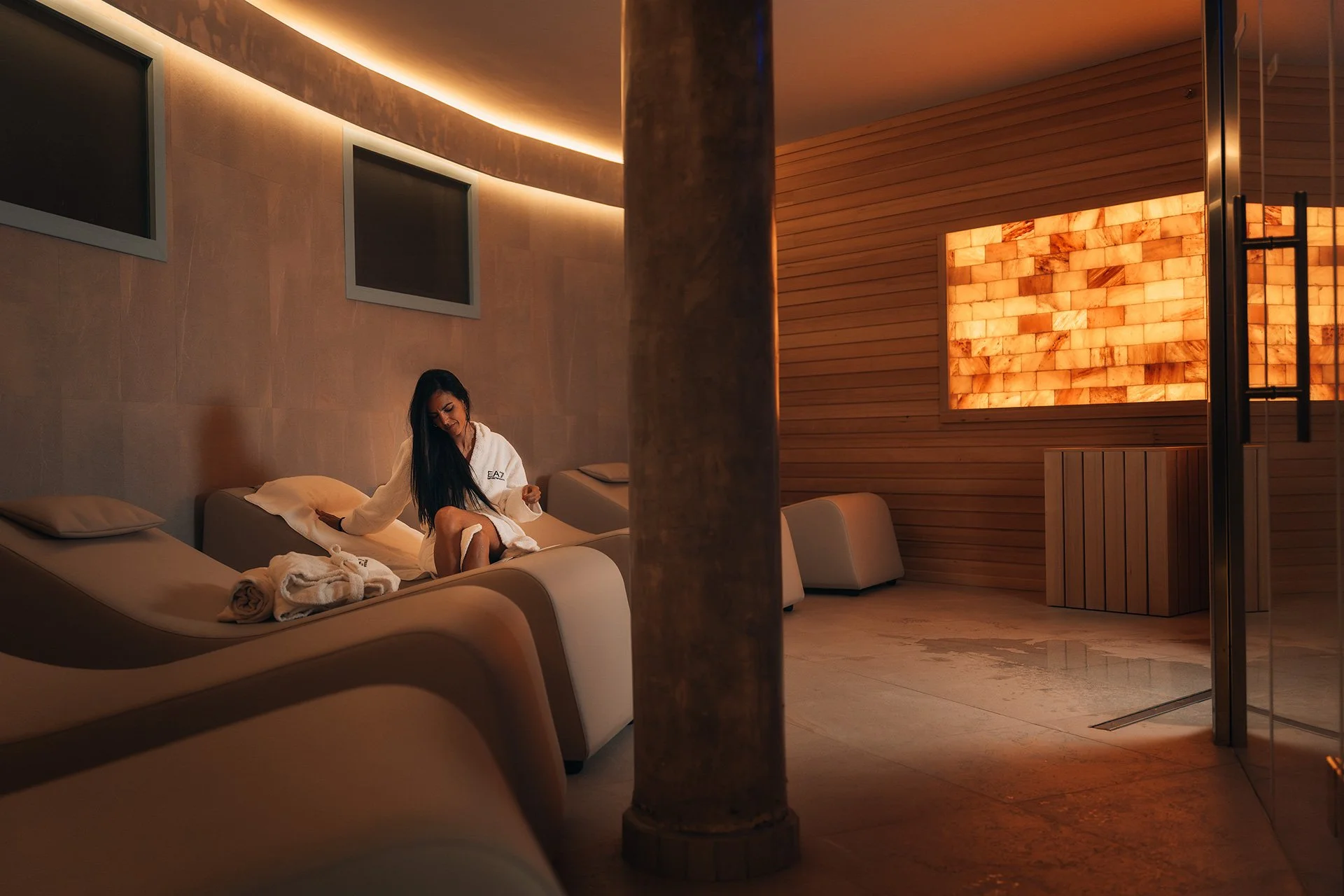 Interno di una spa con pareti in pietra e legno, illuminazione calda, donna seduta su un lettino con accappatoio.