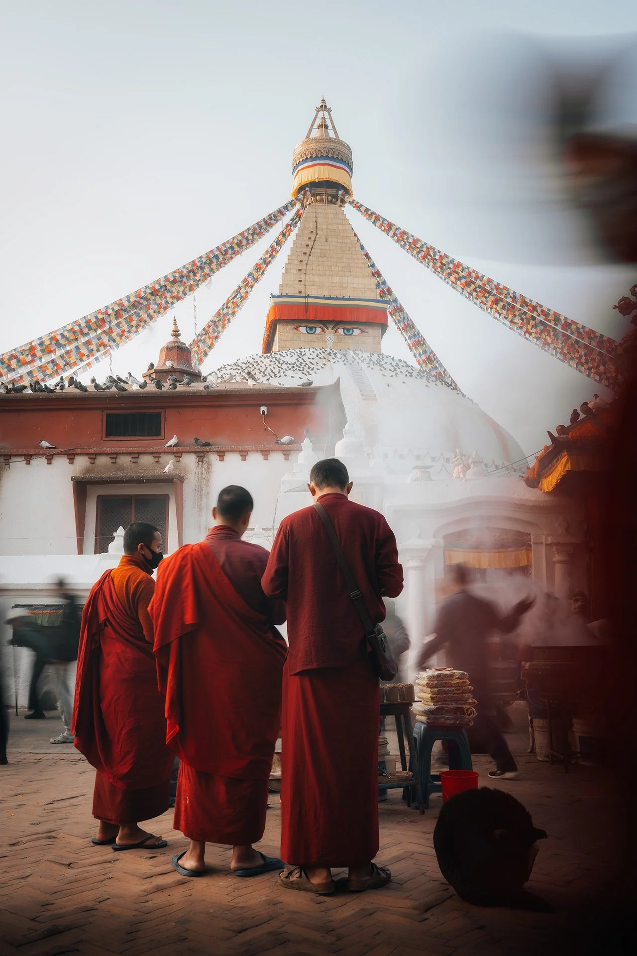 Monaci buddisti vestiti con tuniche rosse che si trovano in una piazza davanti a una grande pagoda con occhi dipinti e drapperie di preghiera appese alla struttura.