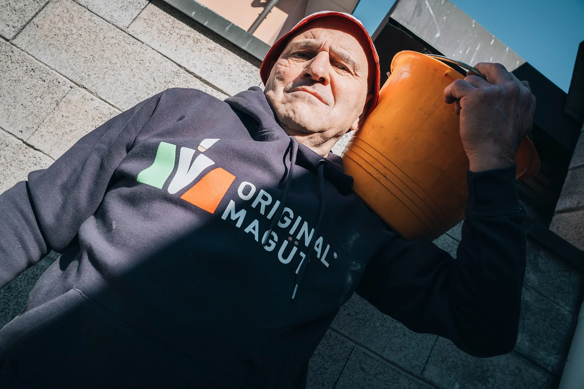 Uomo in uniforme da operaio, indossa un cappello rosso, sdraiato su un marciapiede con un secchio arancione sulla spalla.