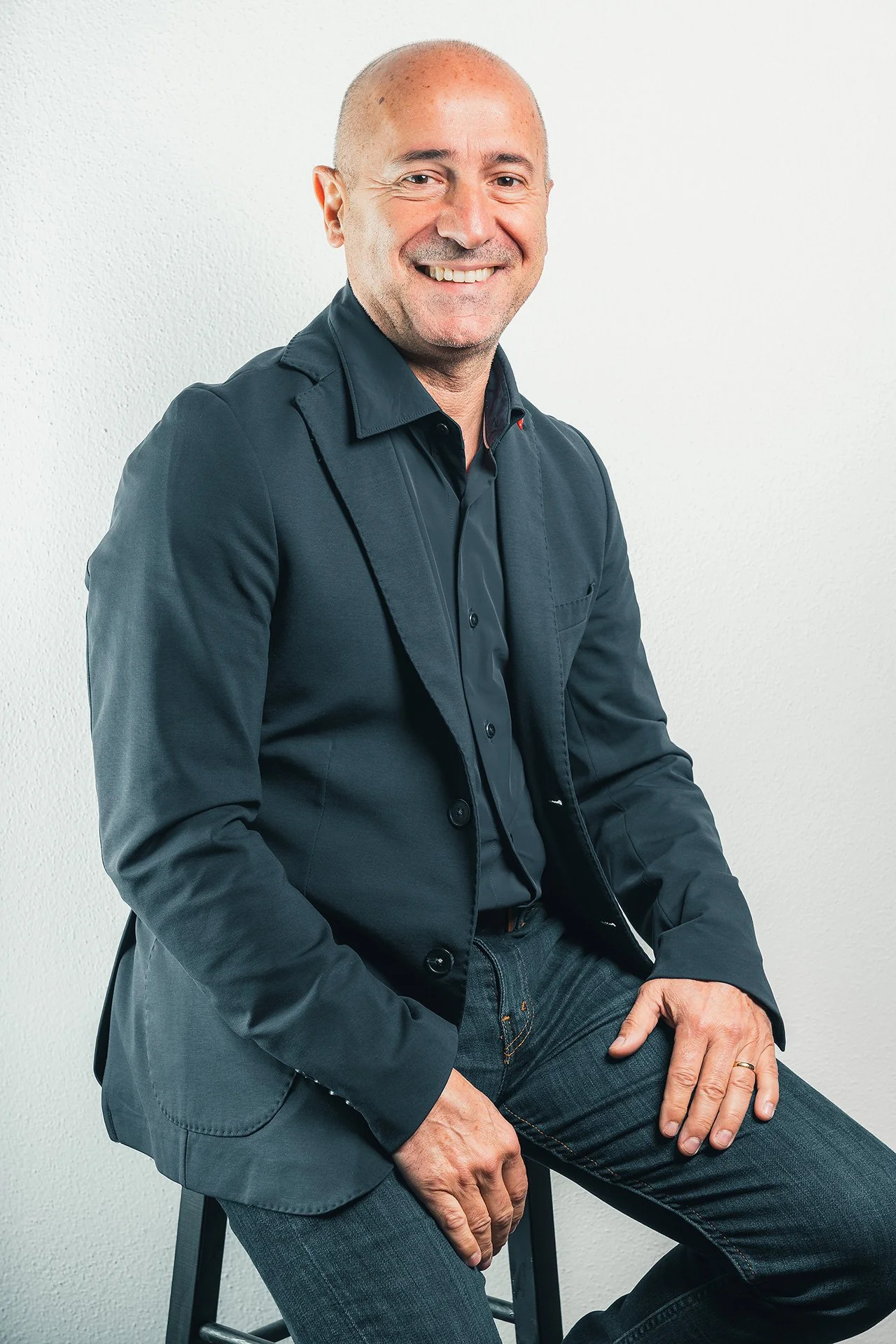 Uomo seduto su uno sgabello, sorridente, con capelli rasati e camicia blu scuro, sfondo bianco.
